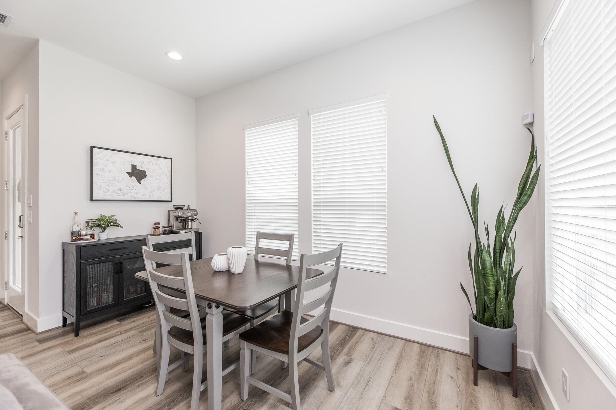 Dining area with a dark wood table, espresso station, and tall snake plant beside bright windows.