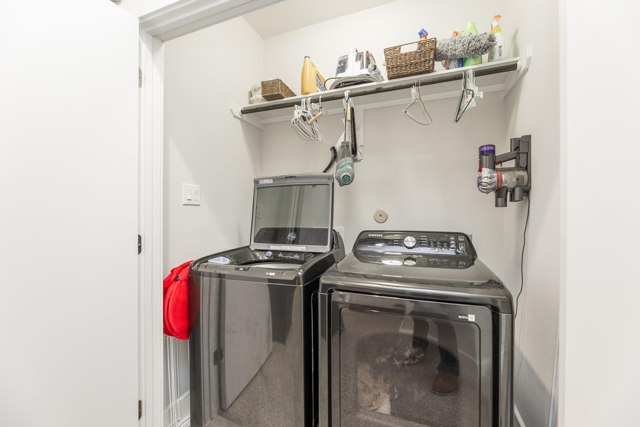 Laundry room with matching dark gray washer and dryer beneath a shelf of cleaning supplies.