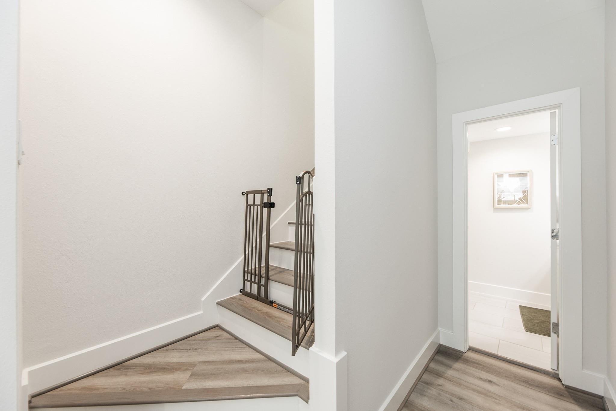 Stairwell with wood-look steps and a dark metal child safety gate beside a bright tiled room.
