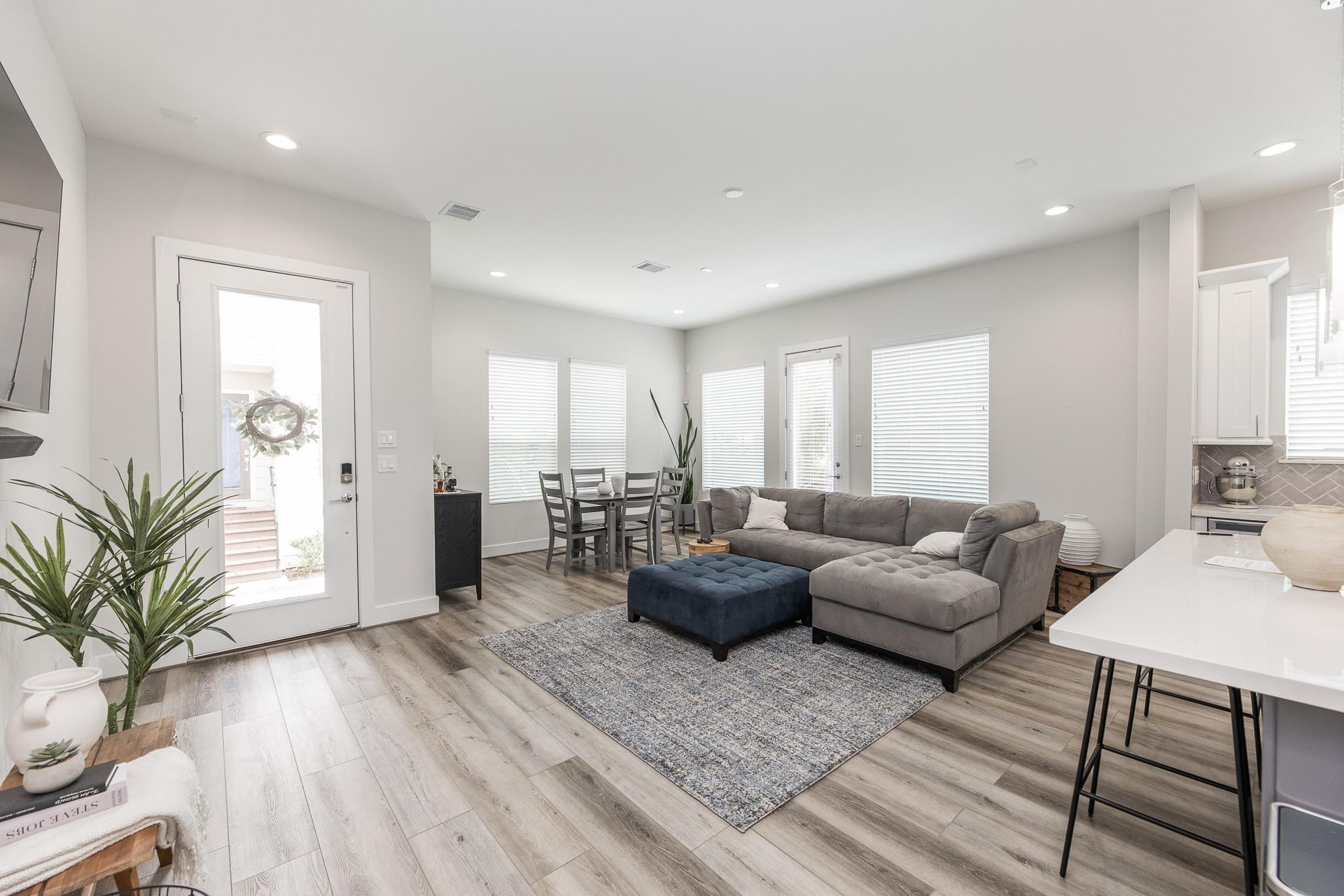 Open-plan living room with gray sectional, blue ottoman, and dining area by bright windows.