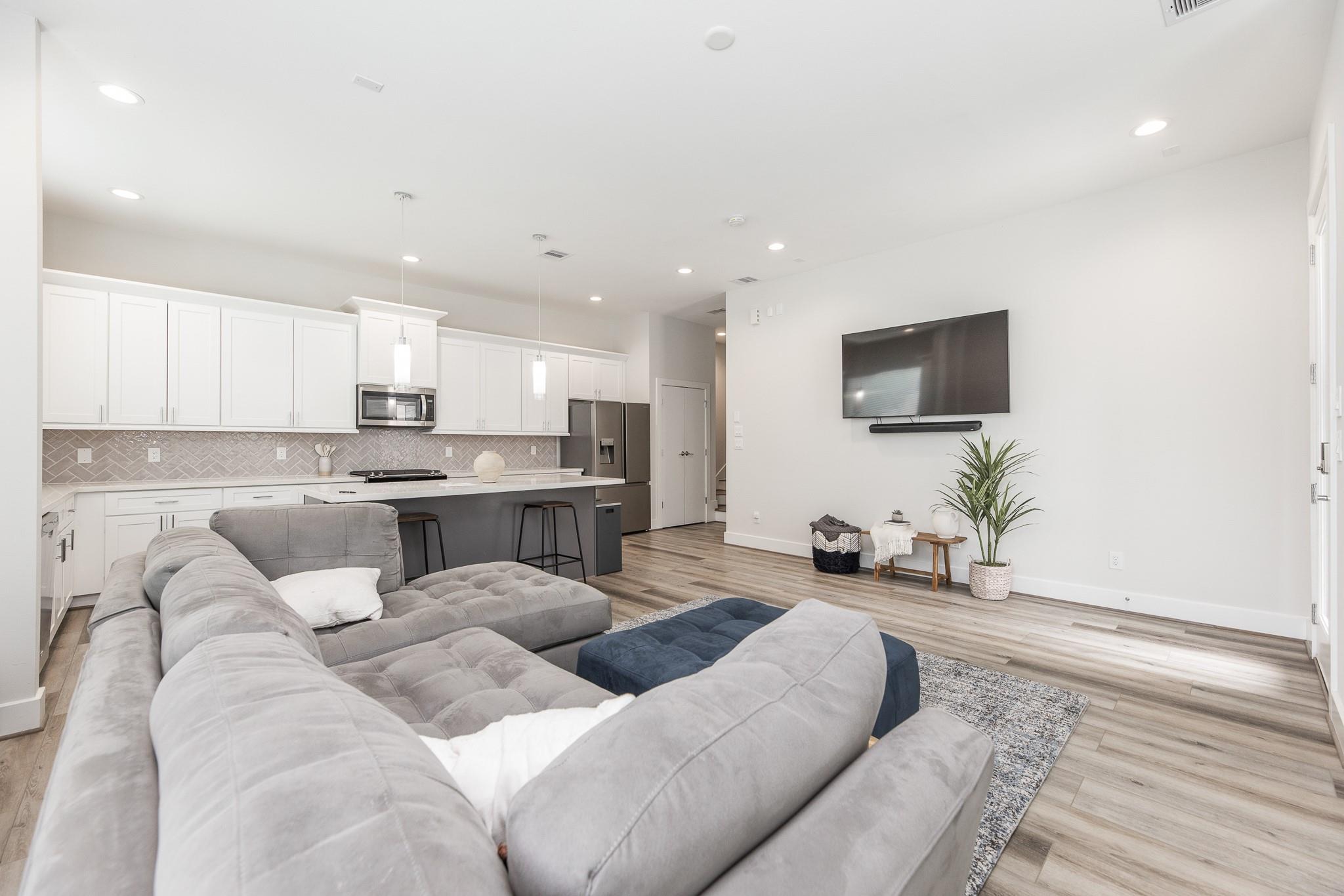 Open-plan living room and kitchen with a large gray sectional, white cabinets, and wall-mounted TV.