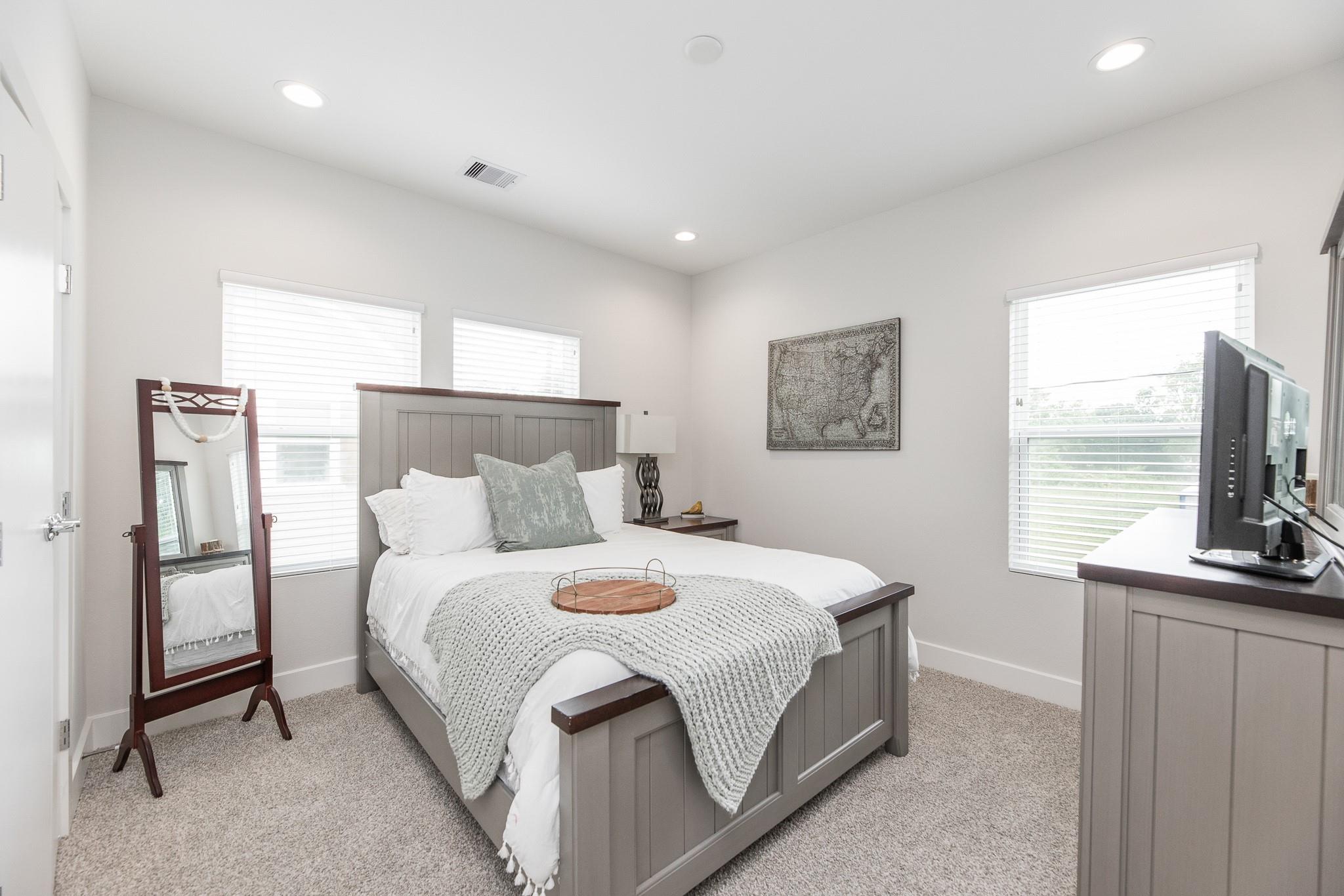 Furnished bedroom with gray paneled bed, white linens, knit blanket, and windows with blinds.