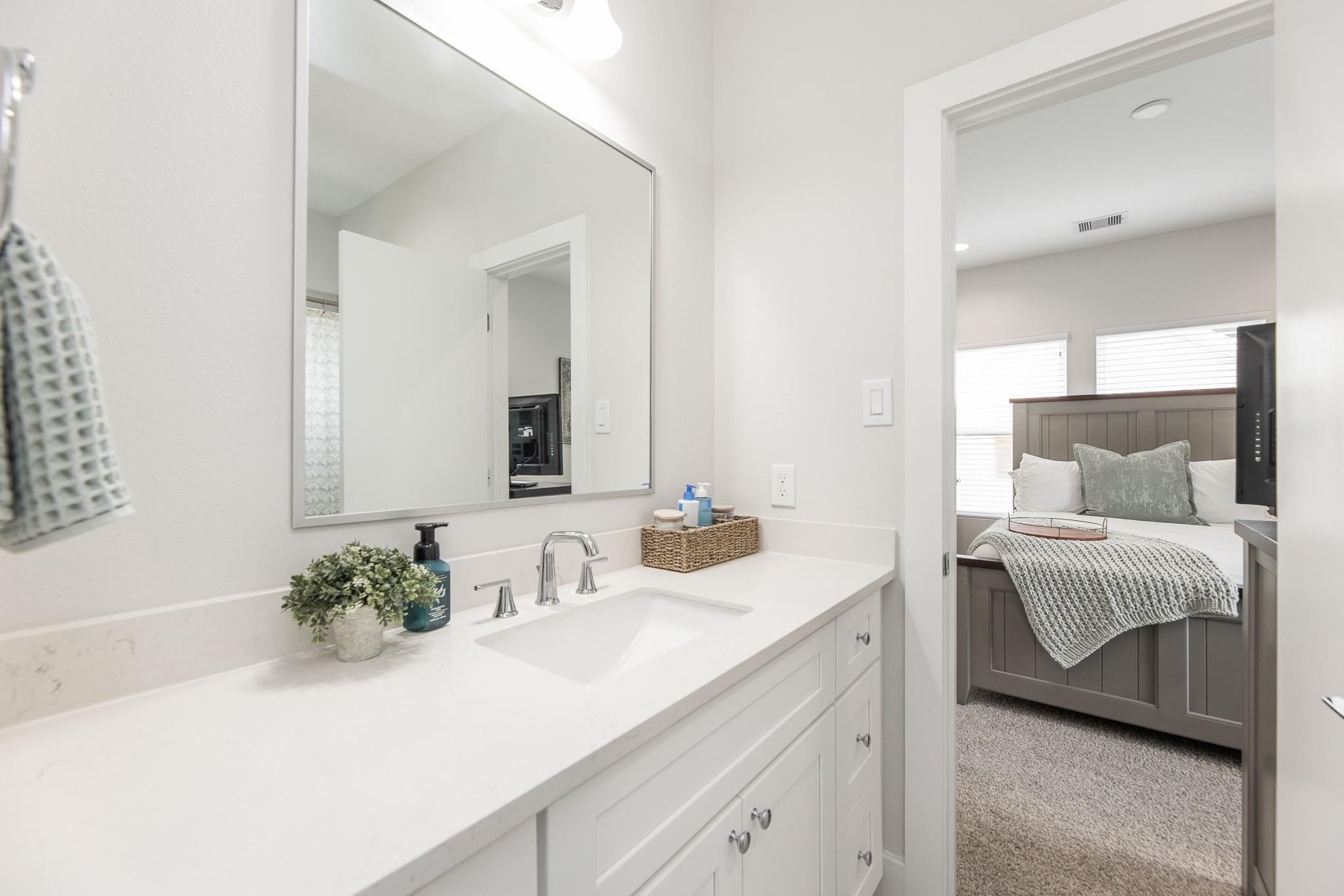 Bathroom vanity with white countertop and large mirror, opening onto a bedroom with a dark paneled bed.