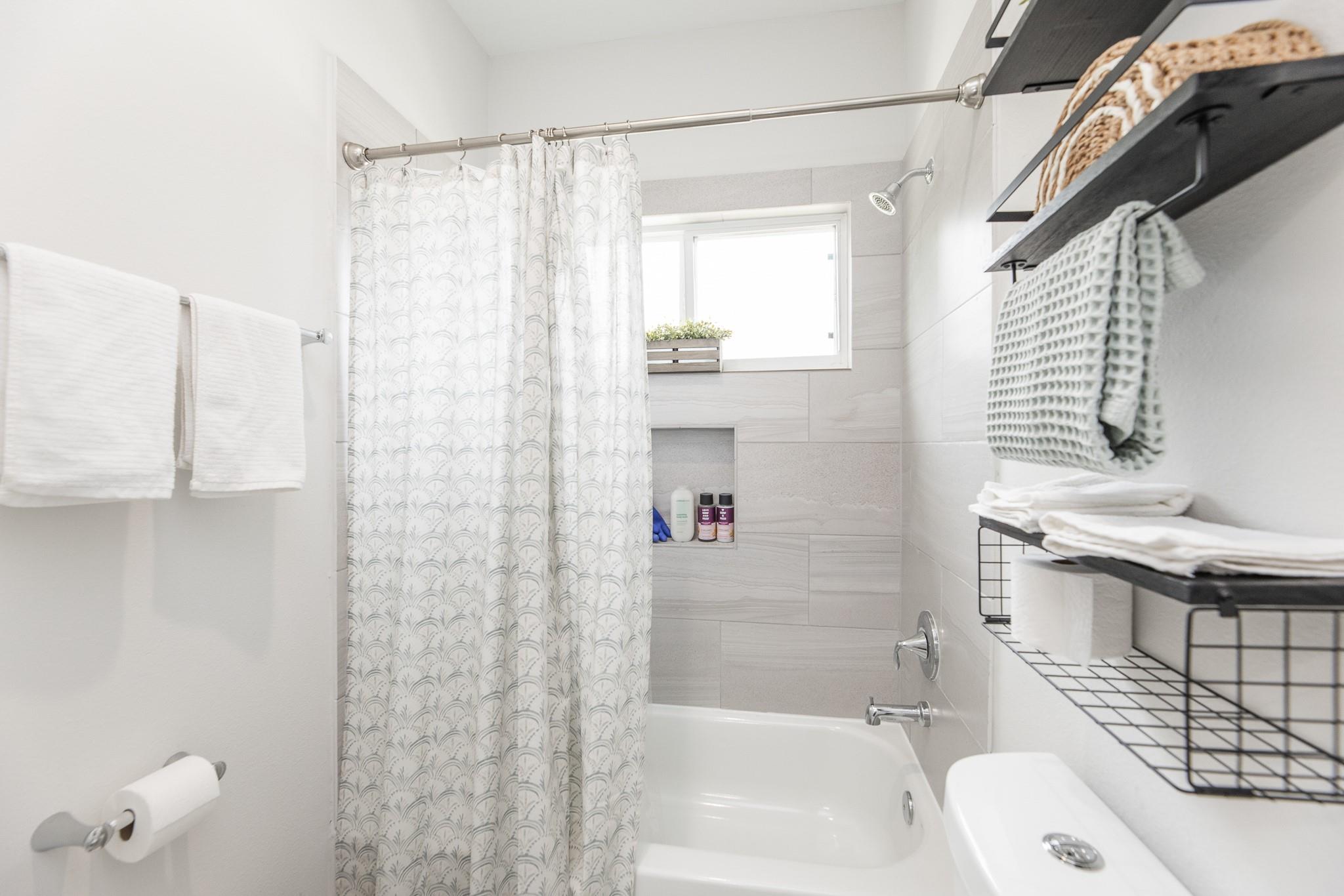 Bright bathroom with a patterned shower curtain, tiled tub surround, and shelves of folded towels.