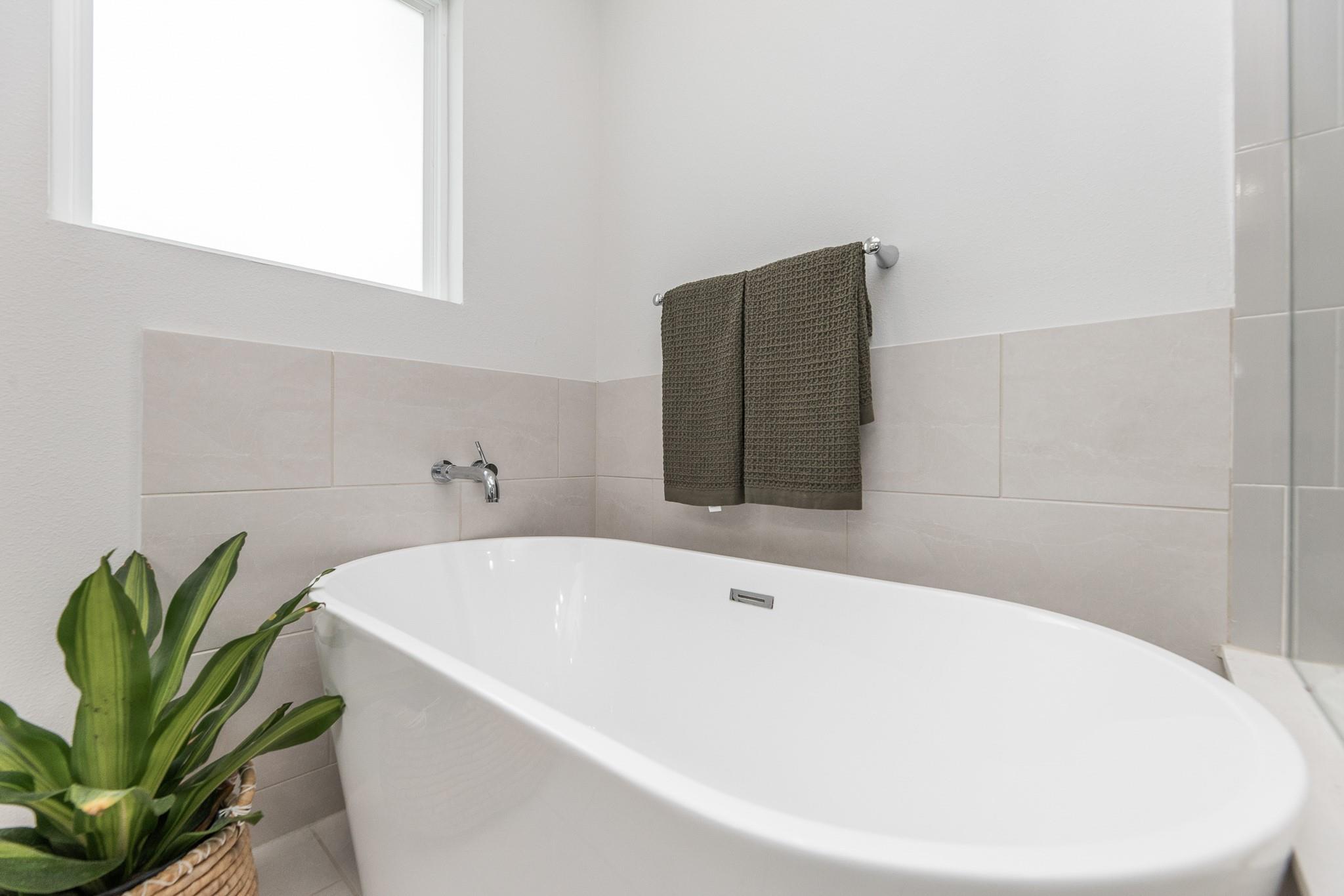 Bathroom with a large white soaking tub, pale tile, dark towel, and leafy potted plant.