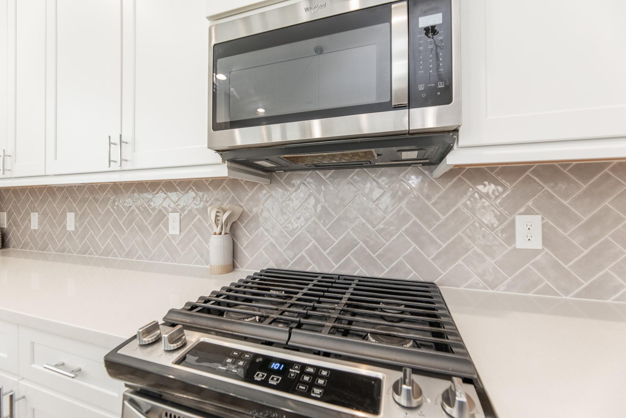 Kitchen range and microwave beneath white cabinets, with a glossy gray herringbone tile backsplash.