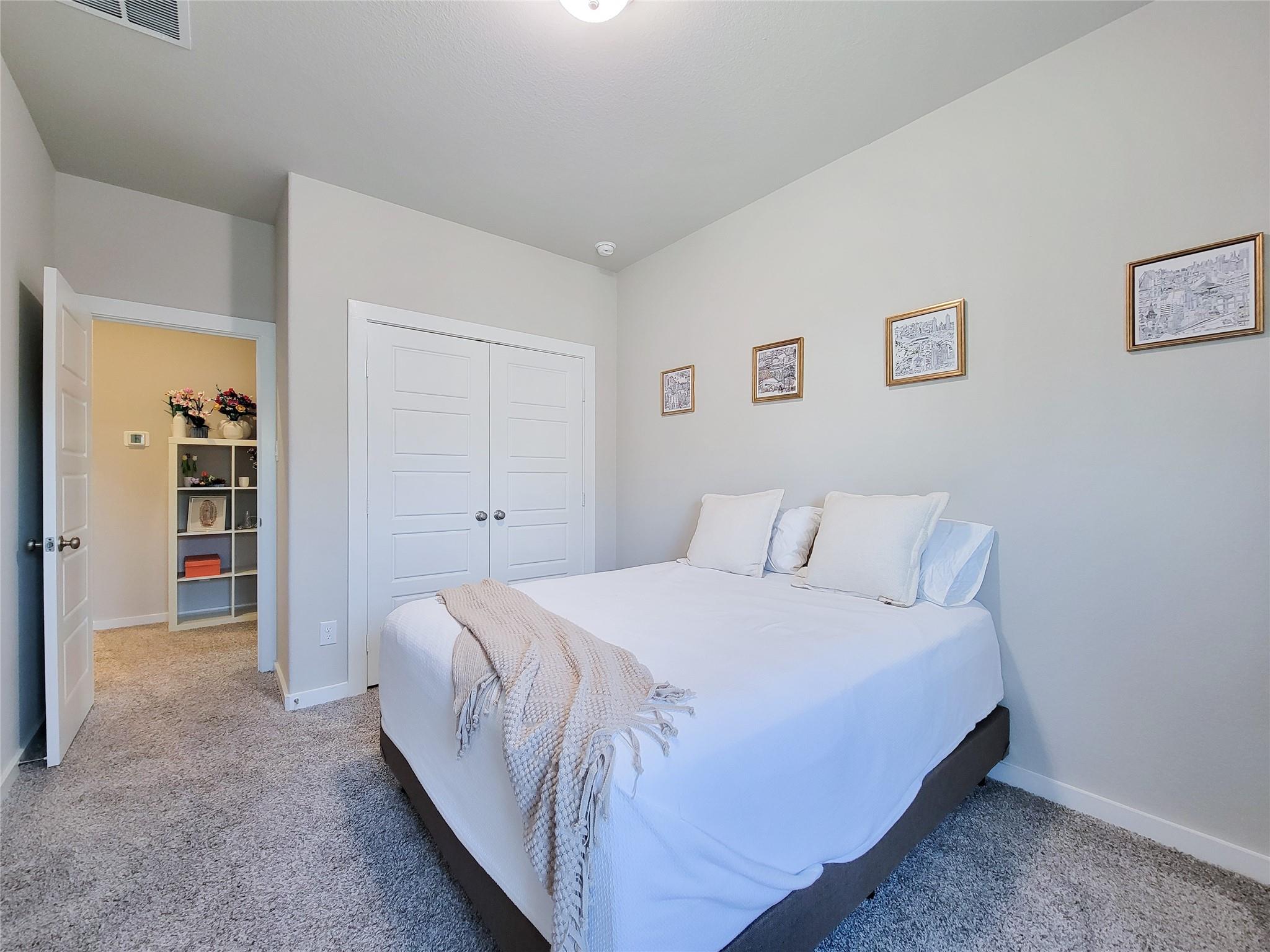 Staged bedroom with a white bed, framed artwork, double closet doors, and an open doorway to a room with shelving.