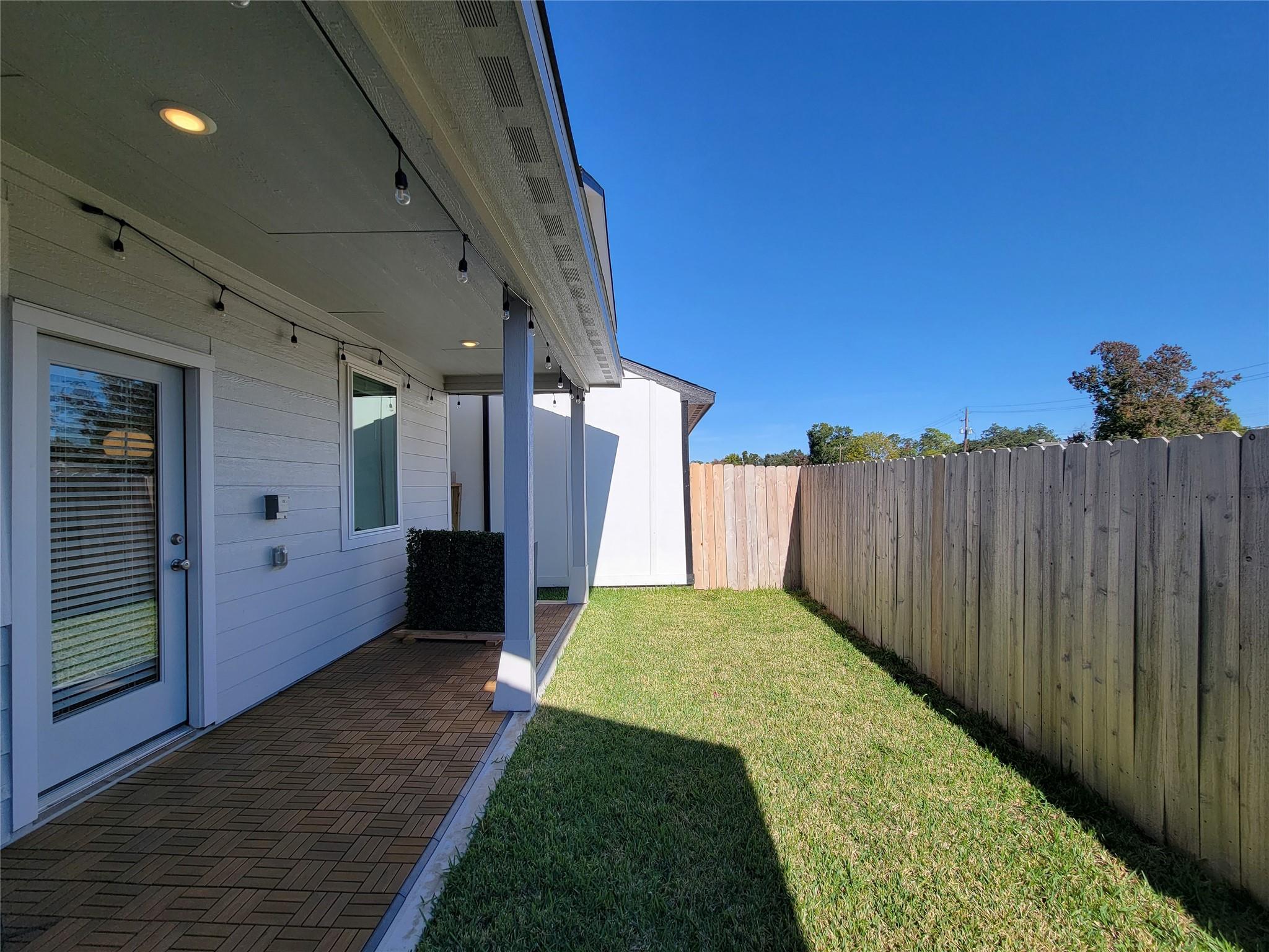 Backyard patio with patterned pavers, a lawn, and a tall wooden privacy fence.