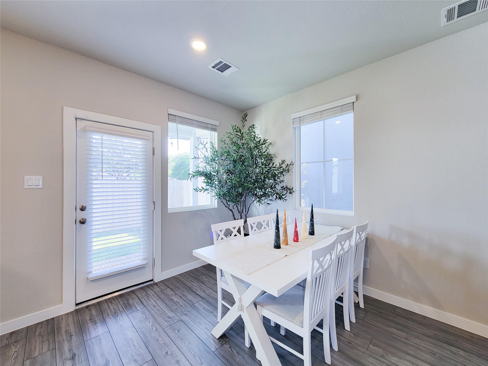 Dining area with a white table and chairs, potted tree, and windows beside a glass door.