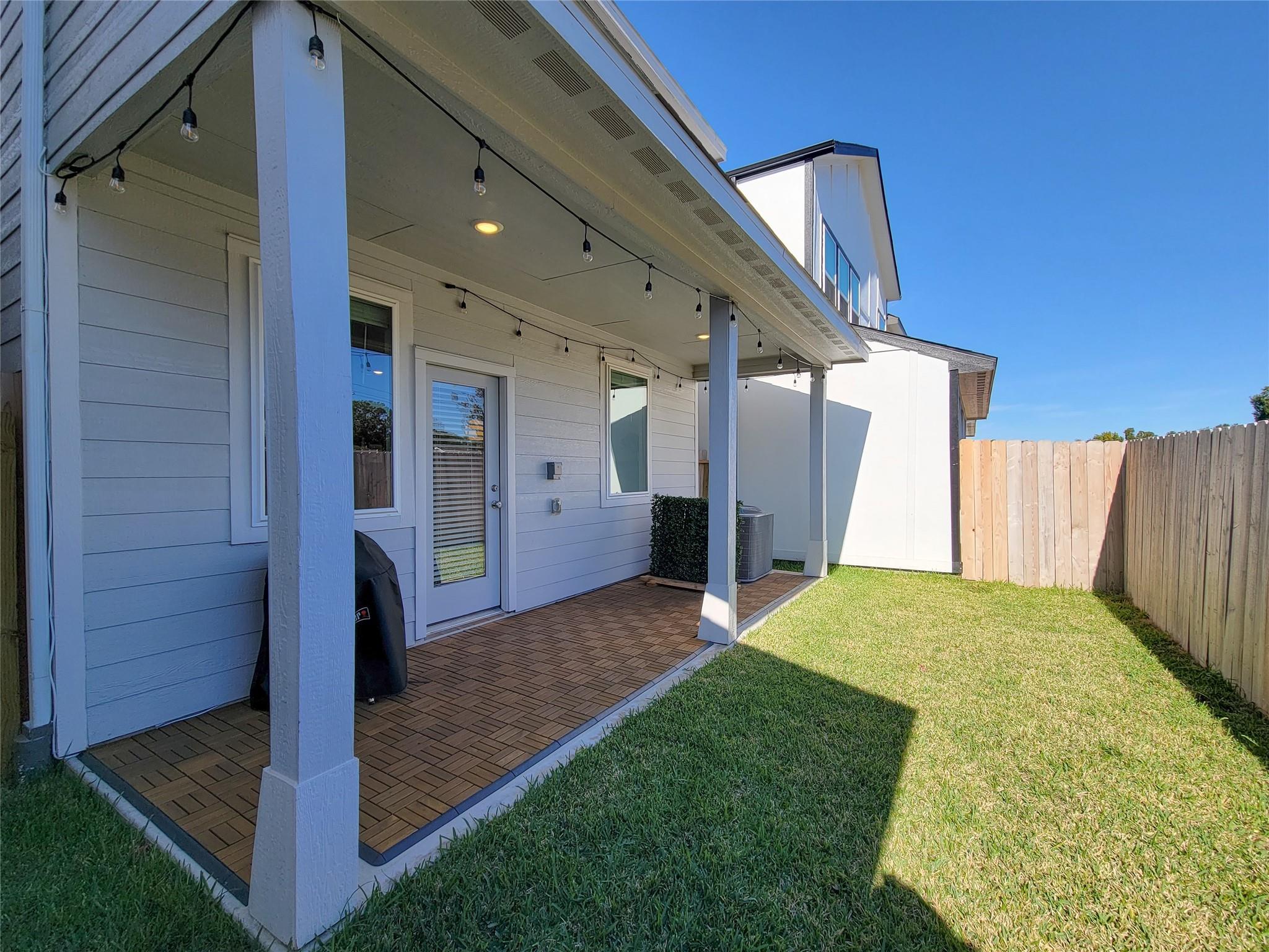 Covered backyard patio with string lights, brown tile flooring, and a fenced lawn.