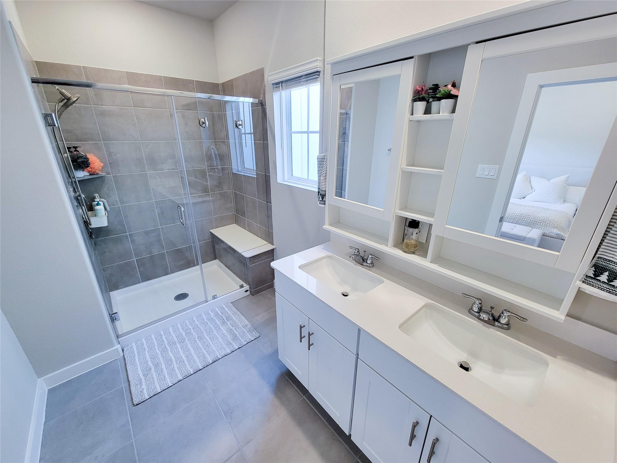 Bathroom with a glass shower, gray tile, built-in bench, and long double-sink vanity.