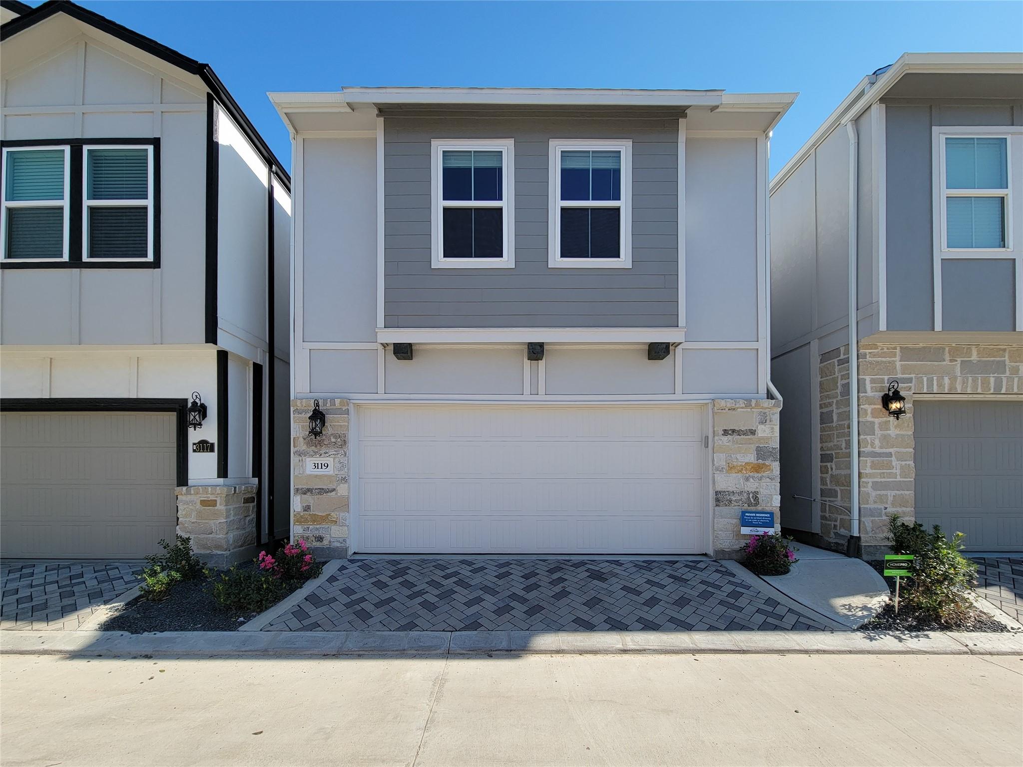 Gray two-story home with white trim, stone accents, and a double garage facing a patterned paver driveway.