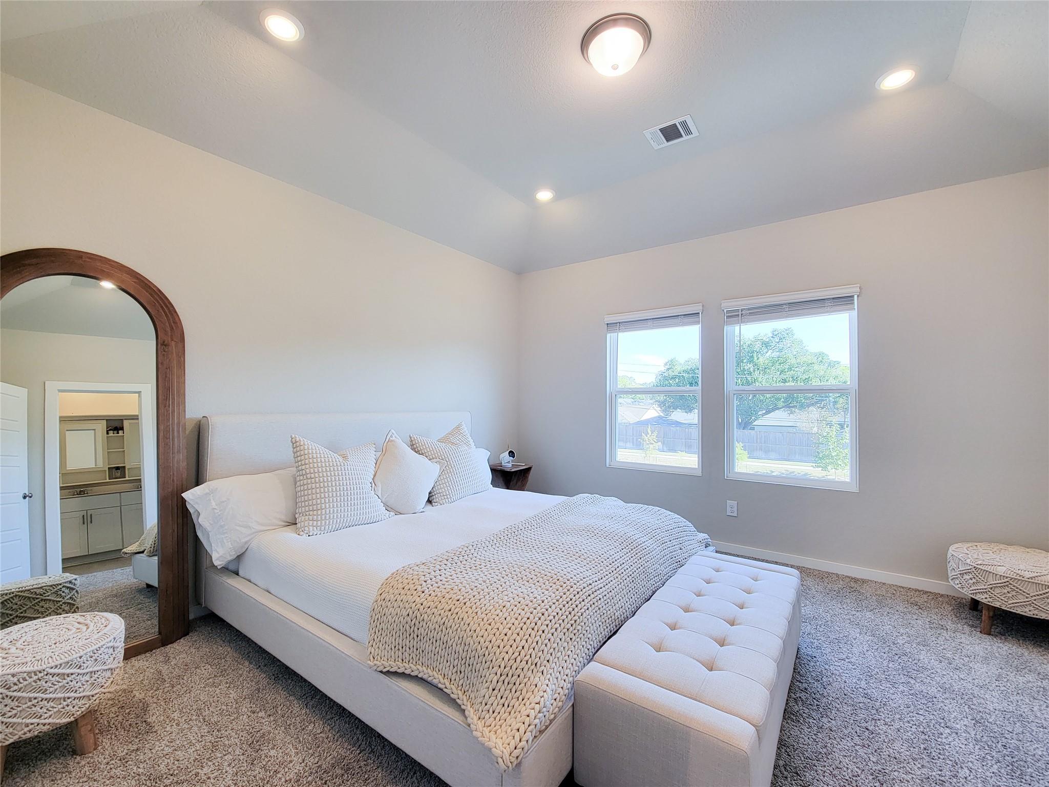 Staged bedroom with a white bed, cream knit blanket, tufted bench, and arched wood-framed mirror.
