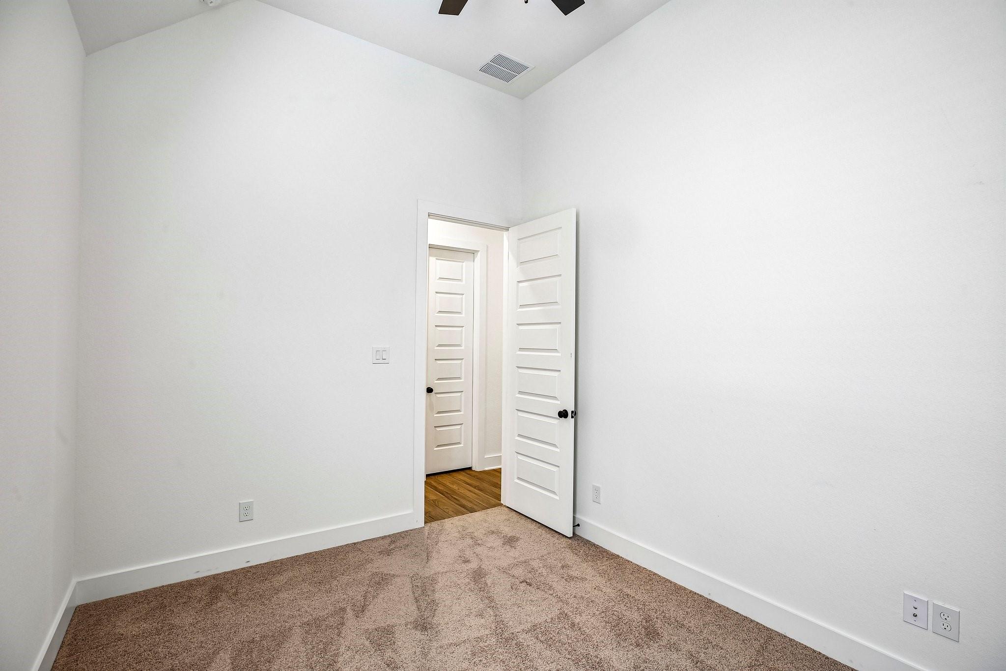 Empty bedroom with beige carpeting and an open door to a wood-floored hallway.