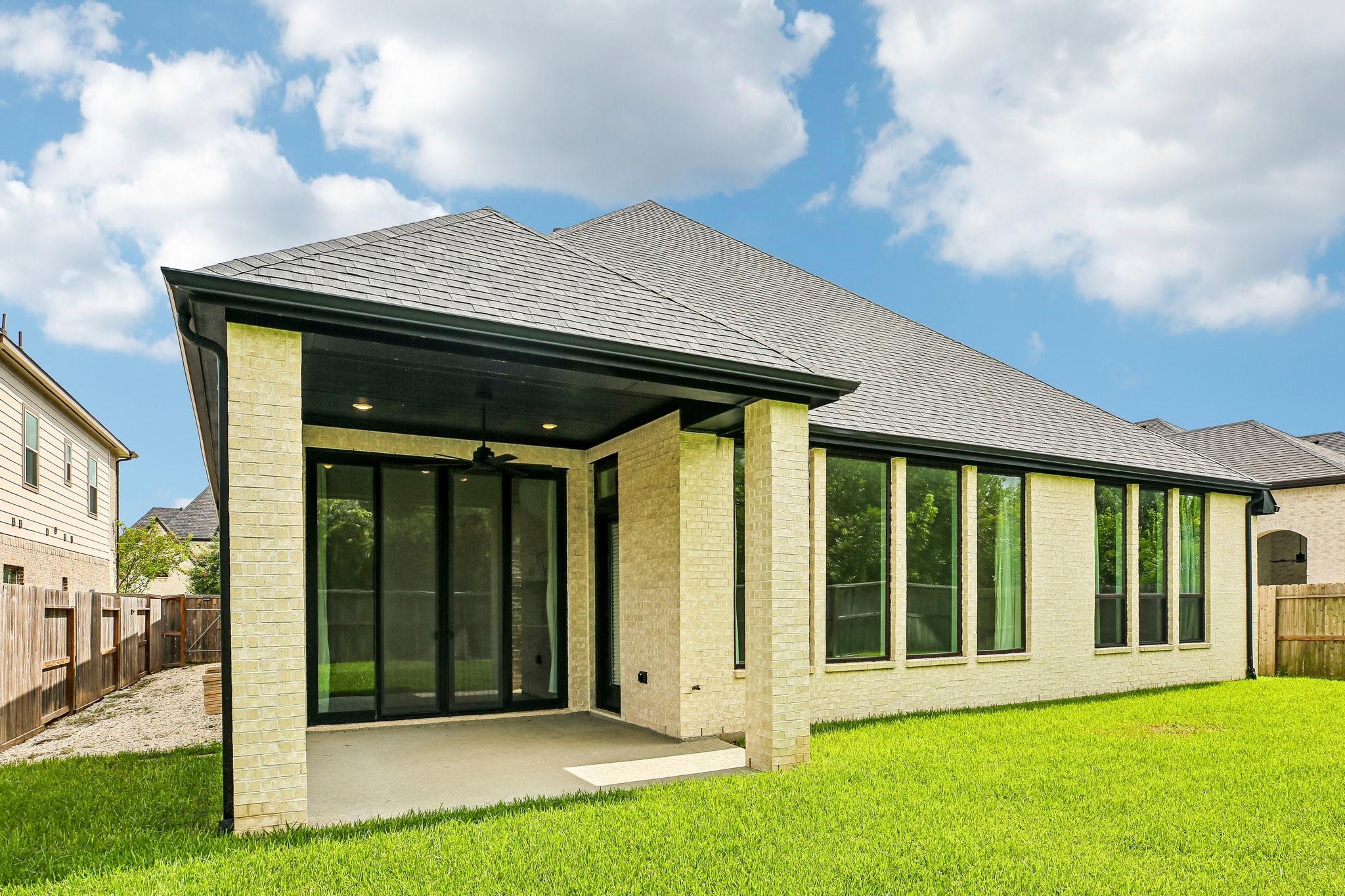 Light brick home with a covered rear patio, dark-framed windows, and a green lawn.