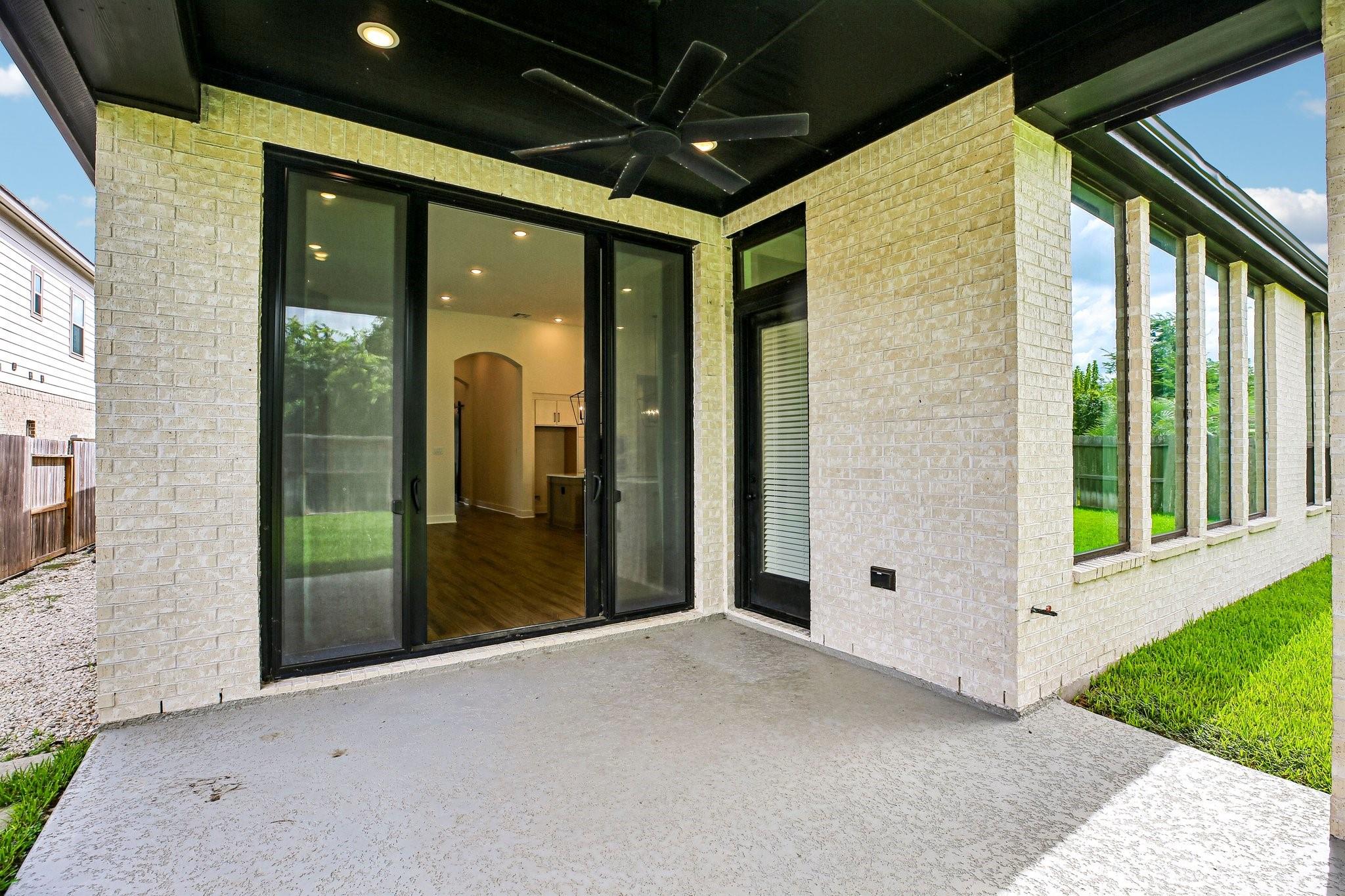 Covered brick patio with a ceiling fan, sliding glass doors, and lawn view.