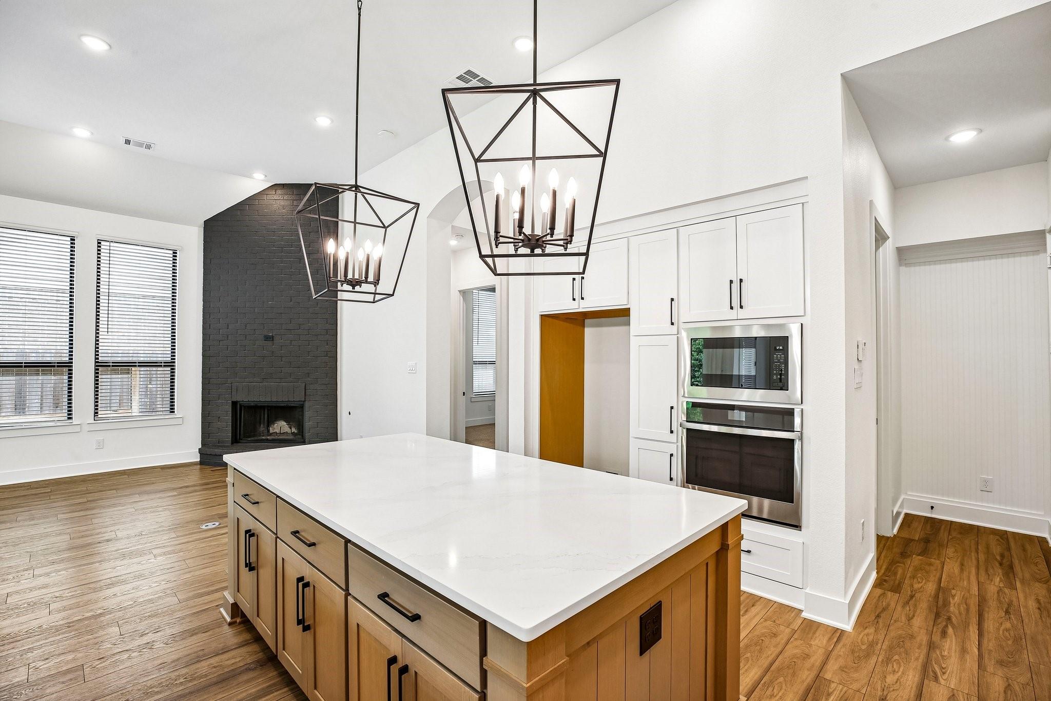 Open kitchen with a large white island, geometric pendant lights, and a dark brick fireplace.