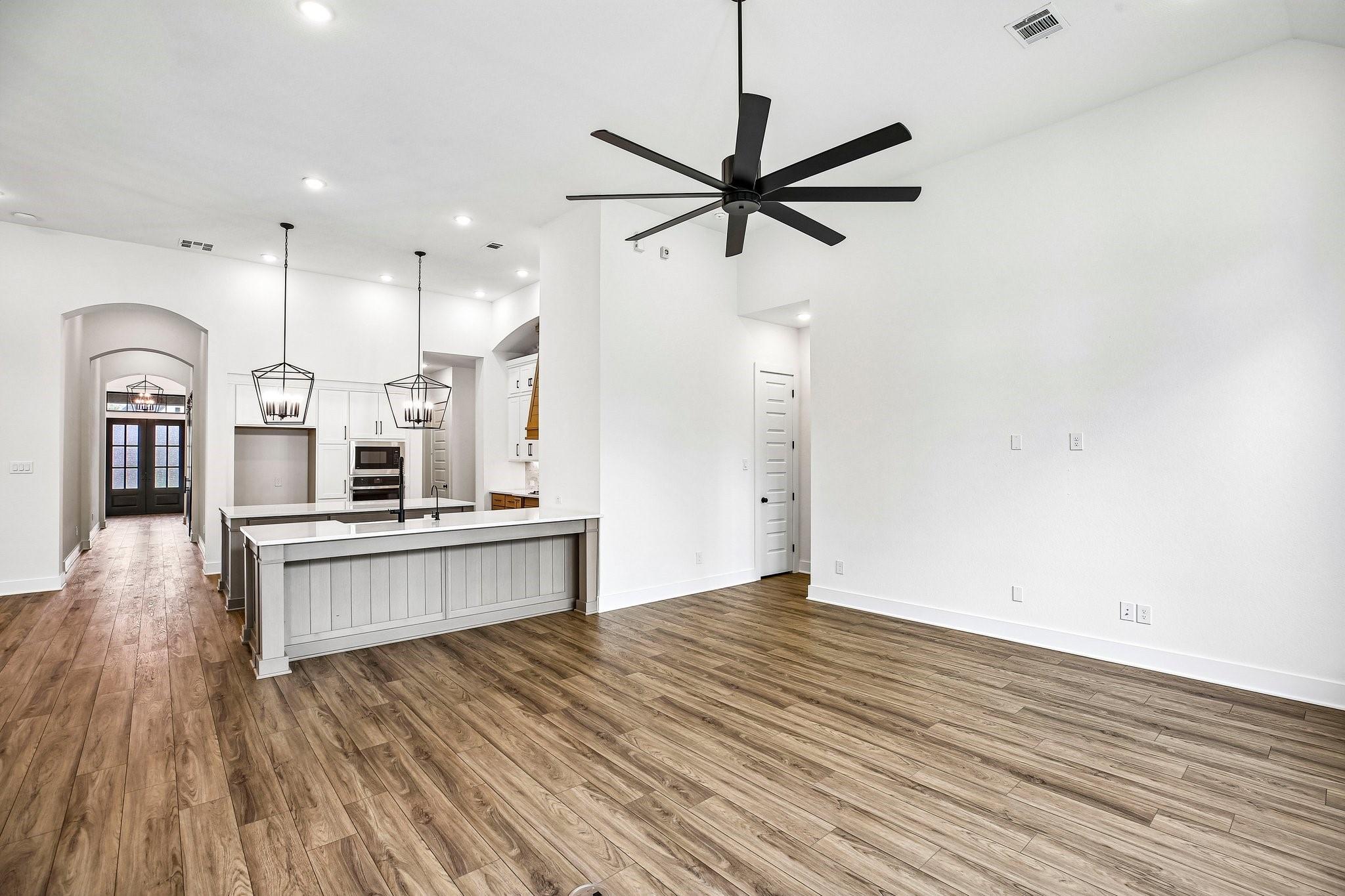 Open-plan kitchen and living area with a large island, wood-look floors, black pendant lights, and ceiling fan.