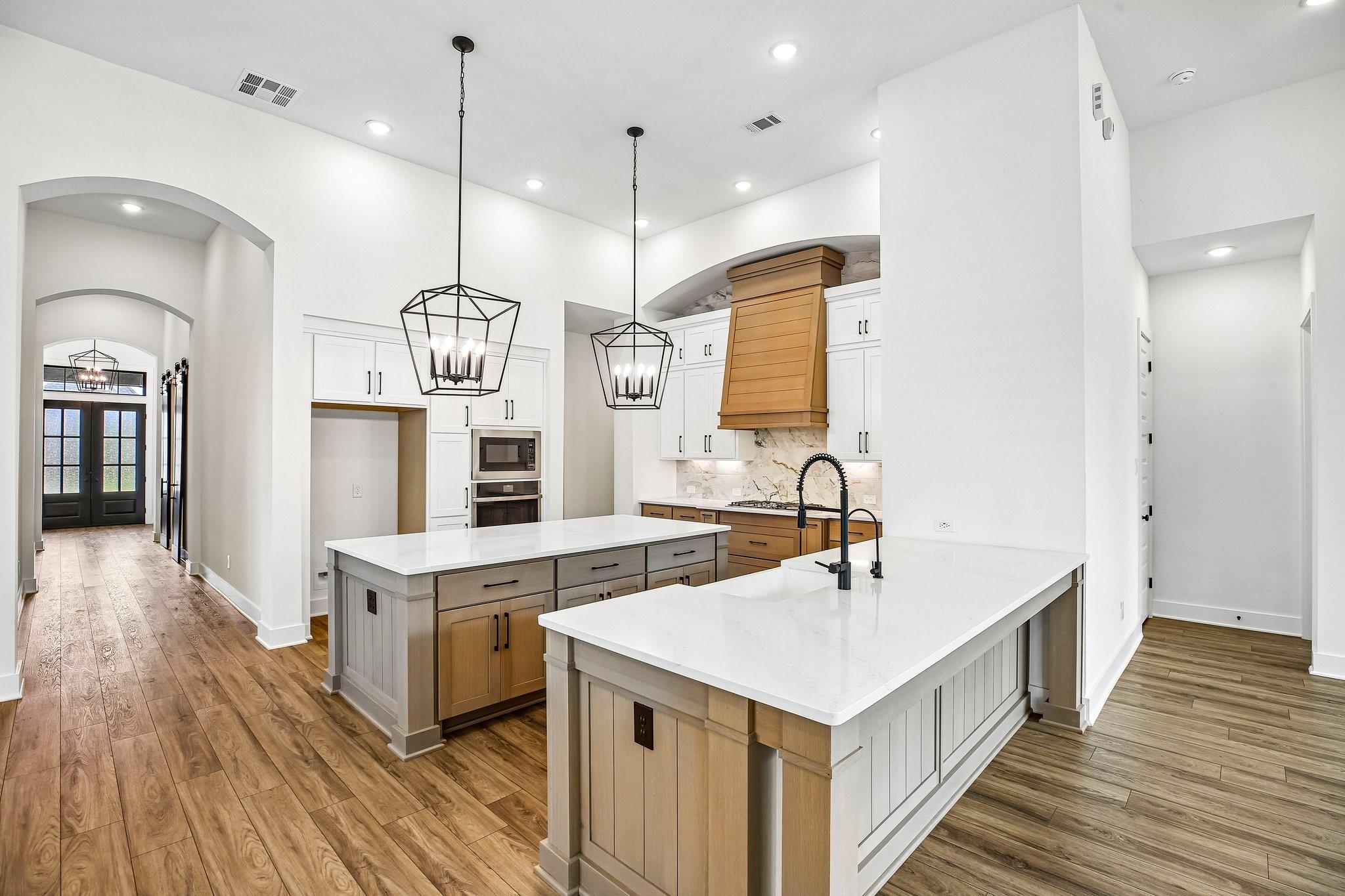 Kitchen with two white-countertop islands, white cabinets, a wood range hood, and geometric pendant lights.