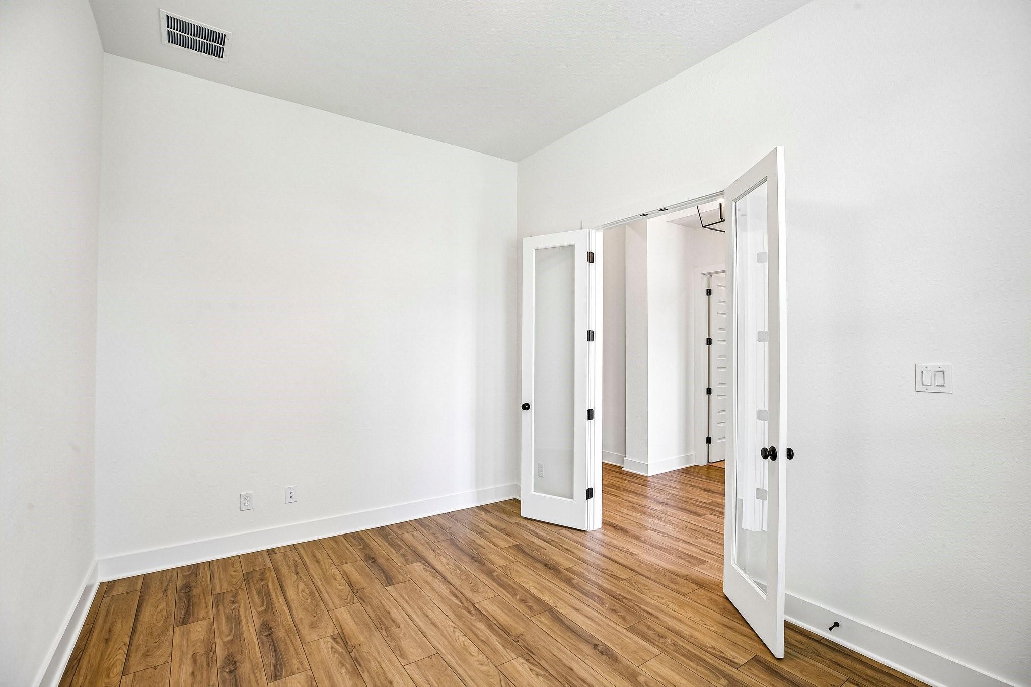 Empty white room with wood-look flooring and open glass-panel French doors to a hallway.