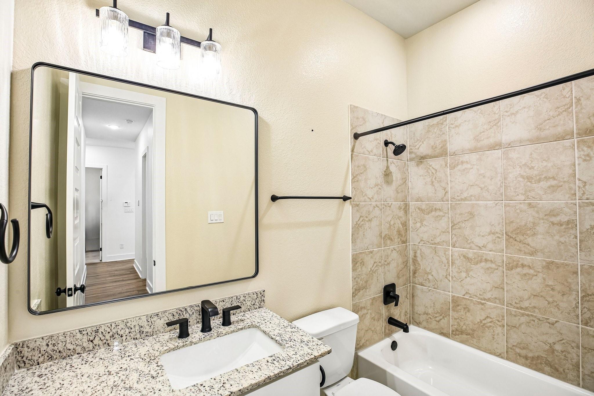 Bathroom with a granite vanity, large mirror, and bathtub with beige tile and black fixtures.