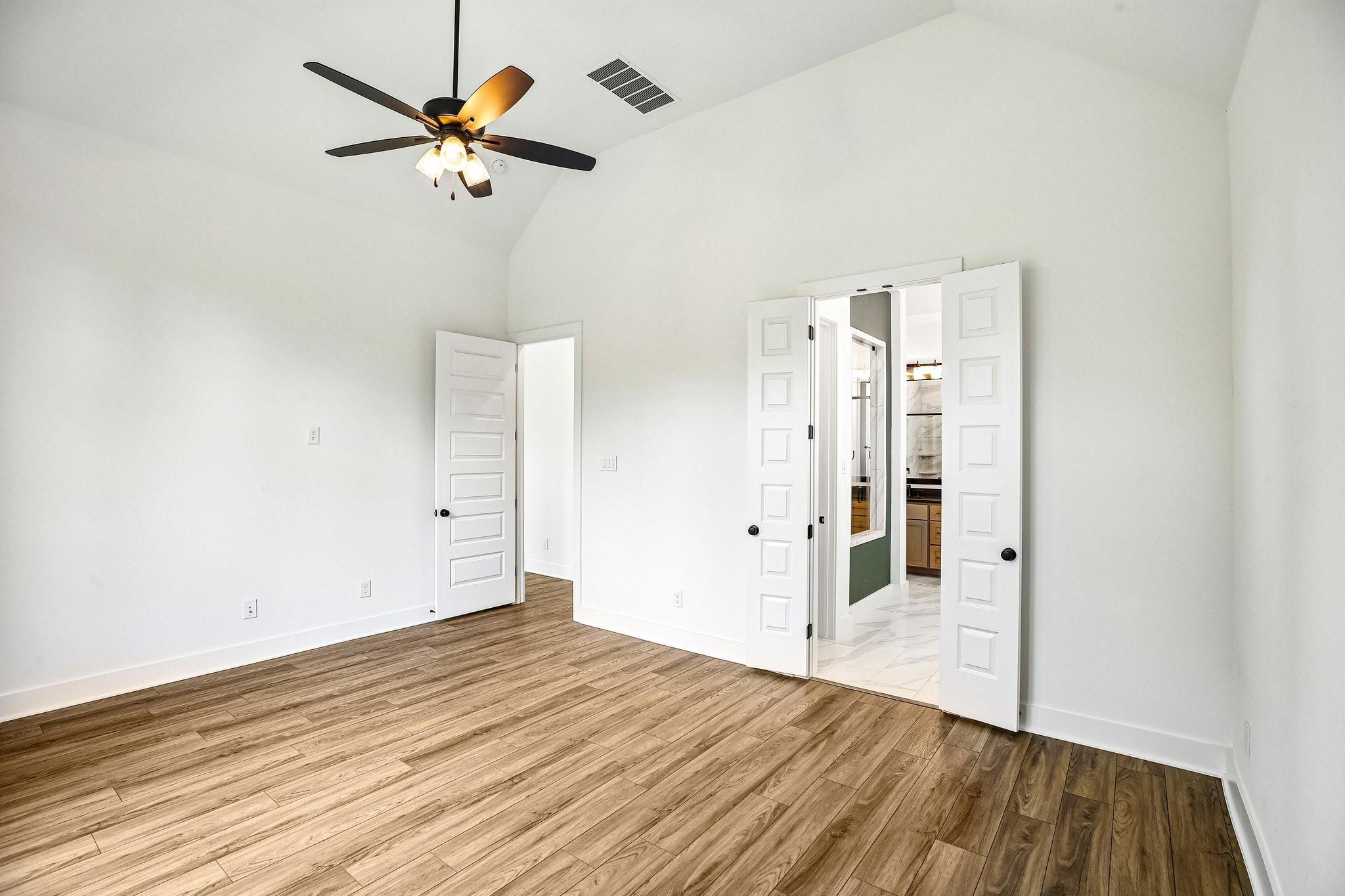 Empty bedroom with wood-look flooring, a ceiling fan, and open double doors to a bathroom.