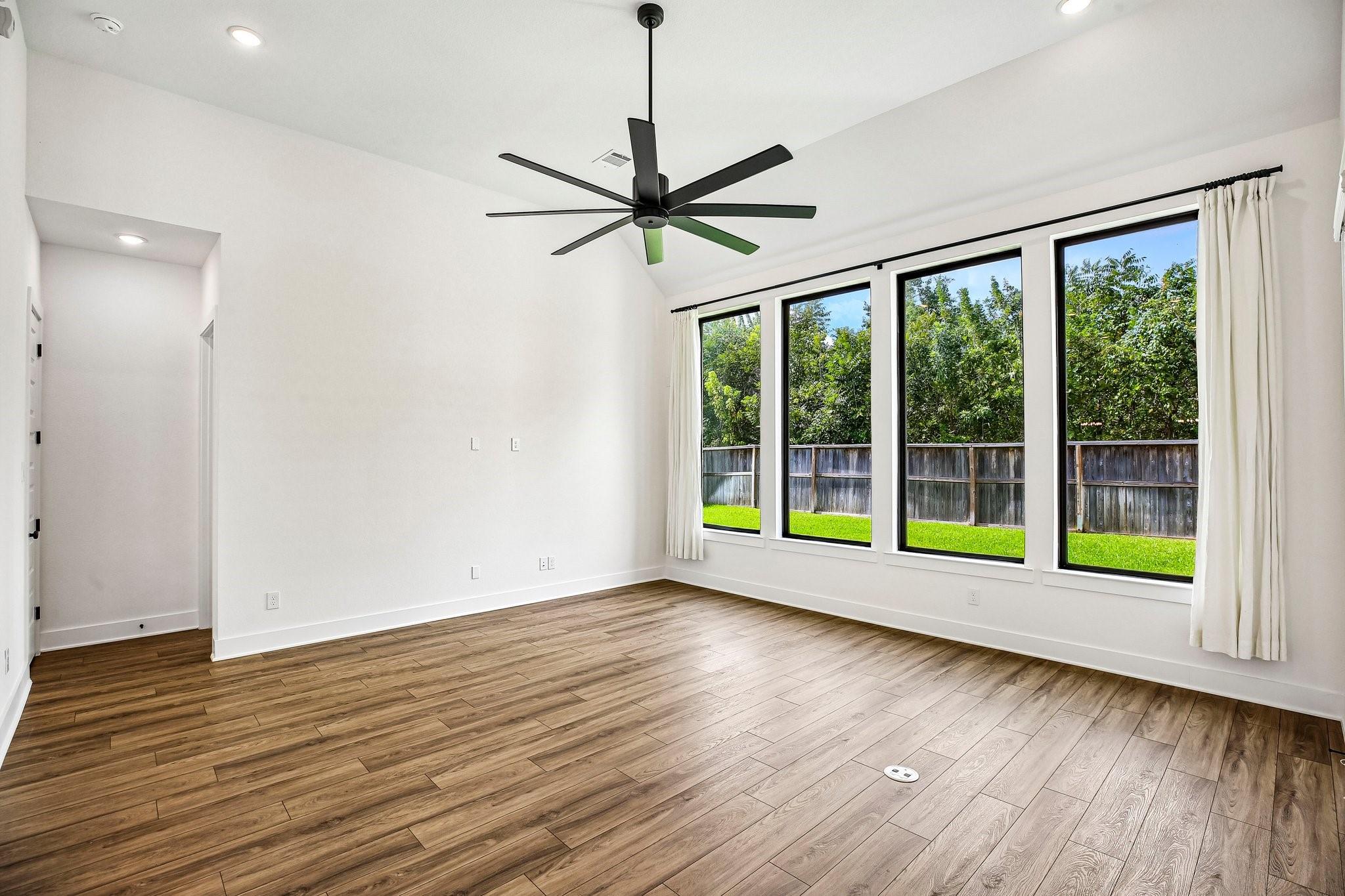 Empty room with wood-look floors, a black ceiling fan, and tall windows facing a fenced green yard.