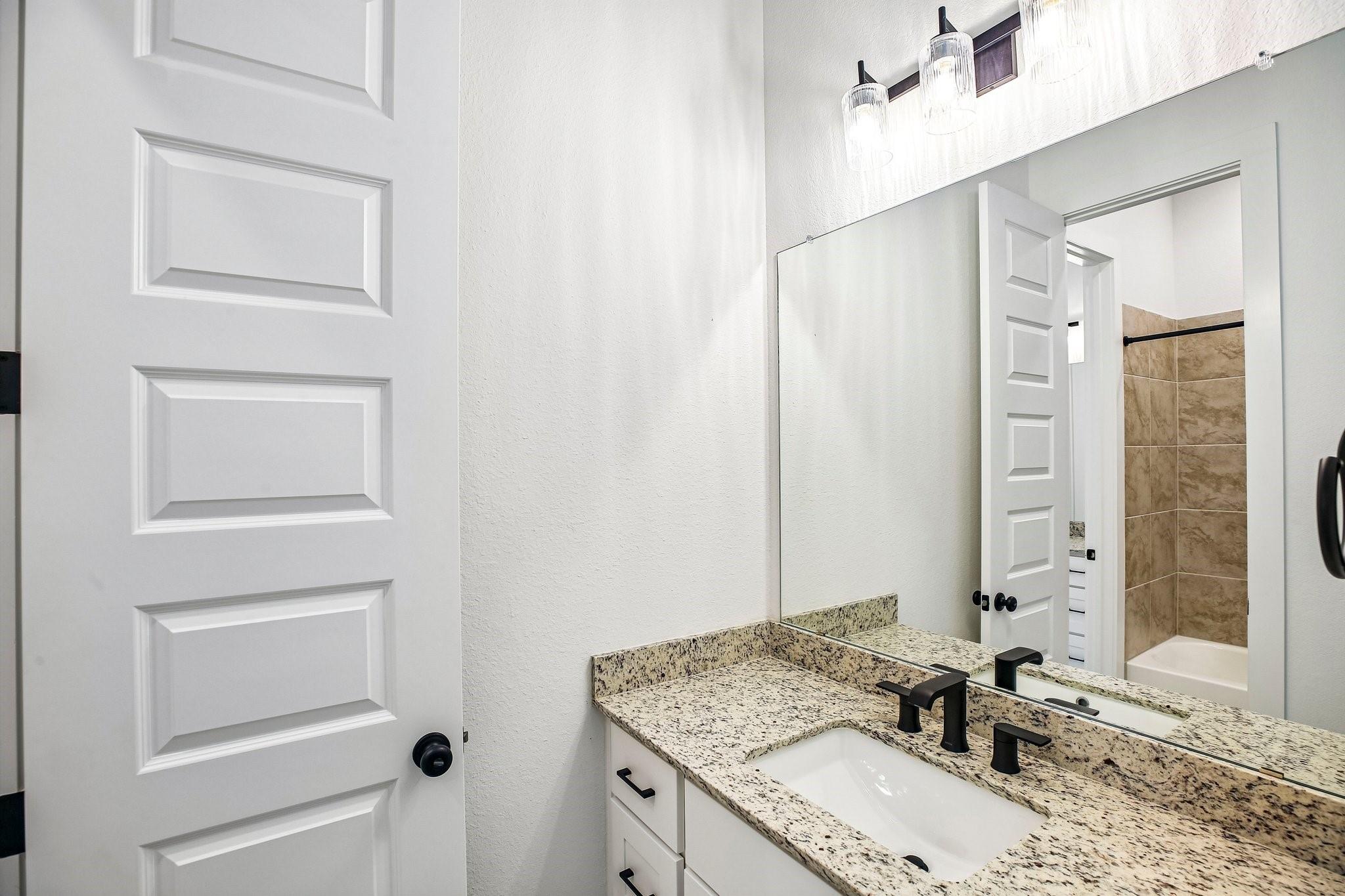 Bathroom vanity with speckled granite counter, black faucet, large mirror, and tan-tiled tub and shower.
