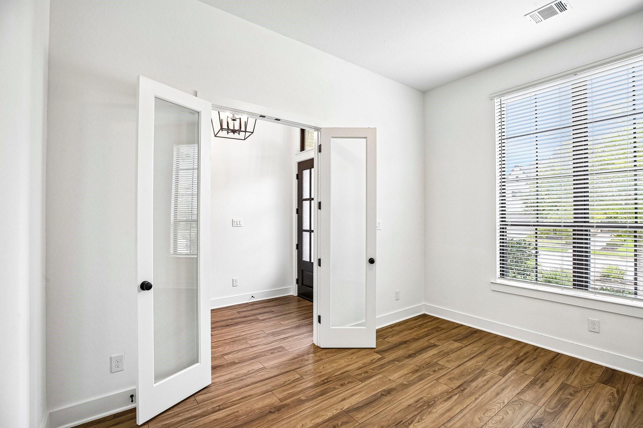 Vacant room with wood-look flooring, large blinded window, and open glass-panel doors to the entry foyer.