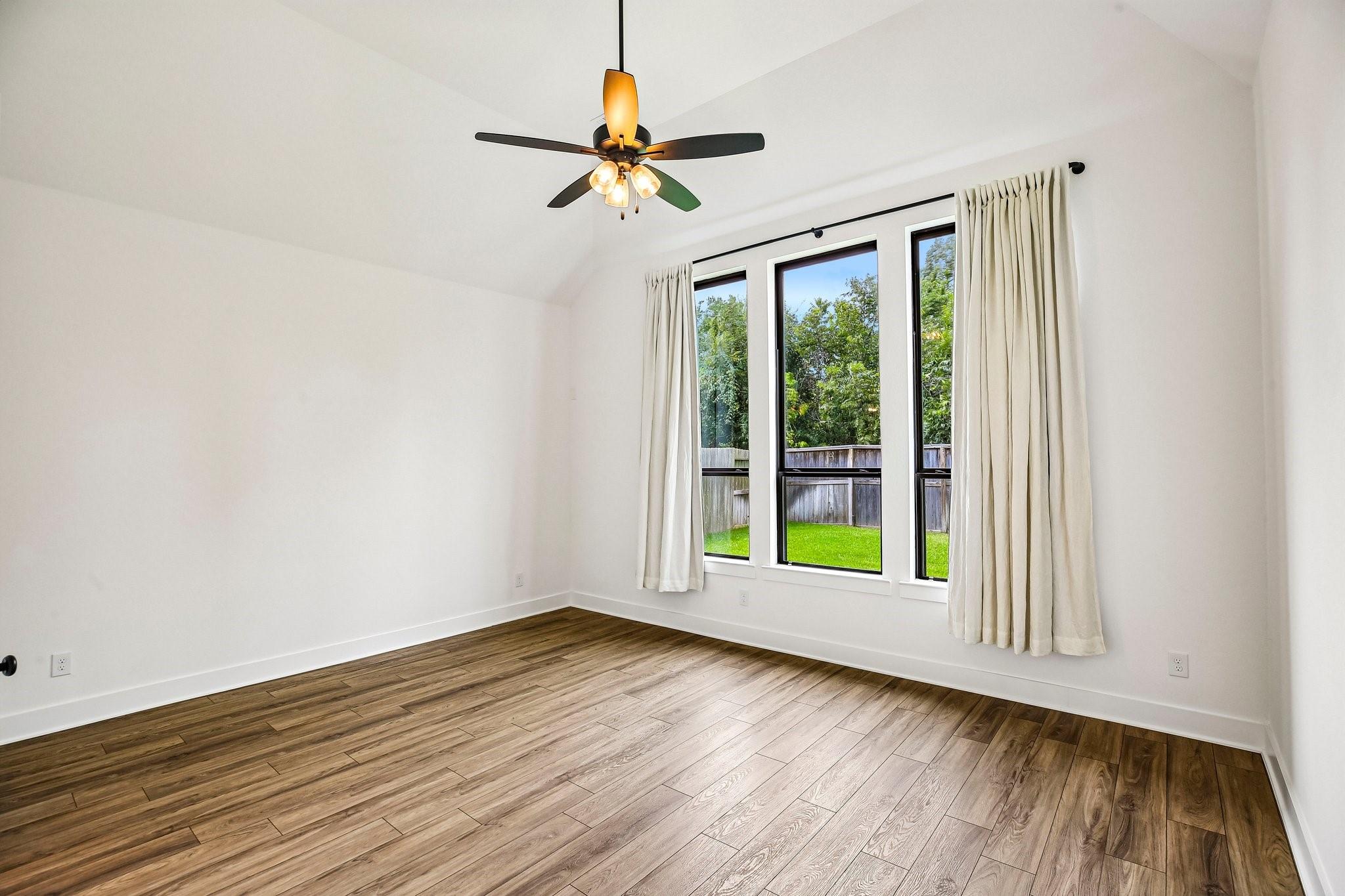Empty room with wood-look flooring, a ceiling fan, and tall windows overlooking a fenced yard.