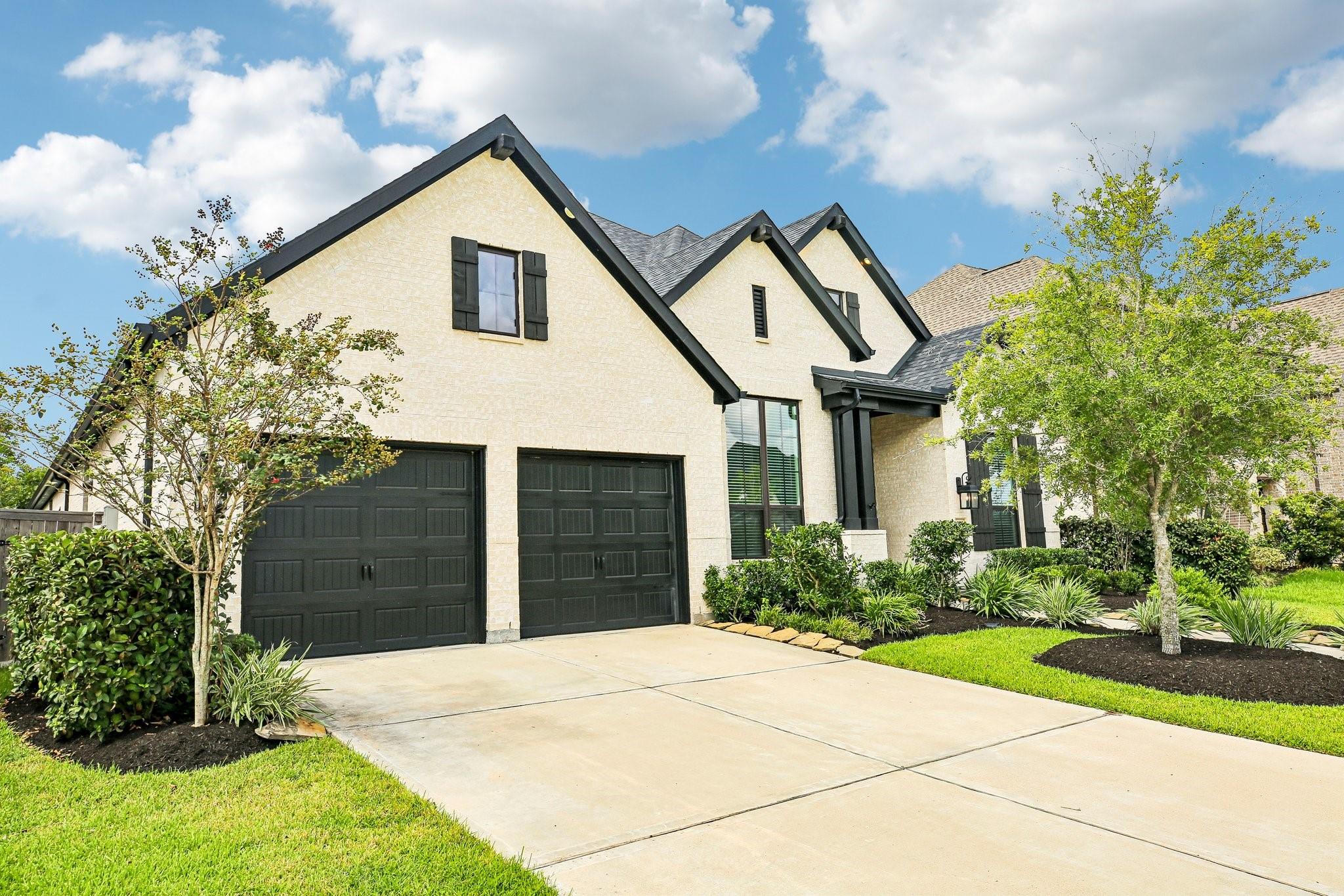 Light brick house with black double garage doors, a wide driveway, and landscaped front yard.