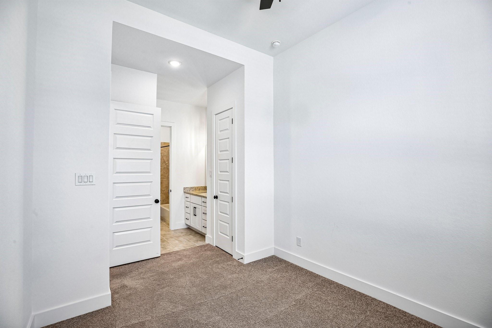 Empty carpeted bedroom with white walls and open doors to a bathroom vanity and tiled shower.