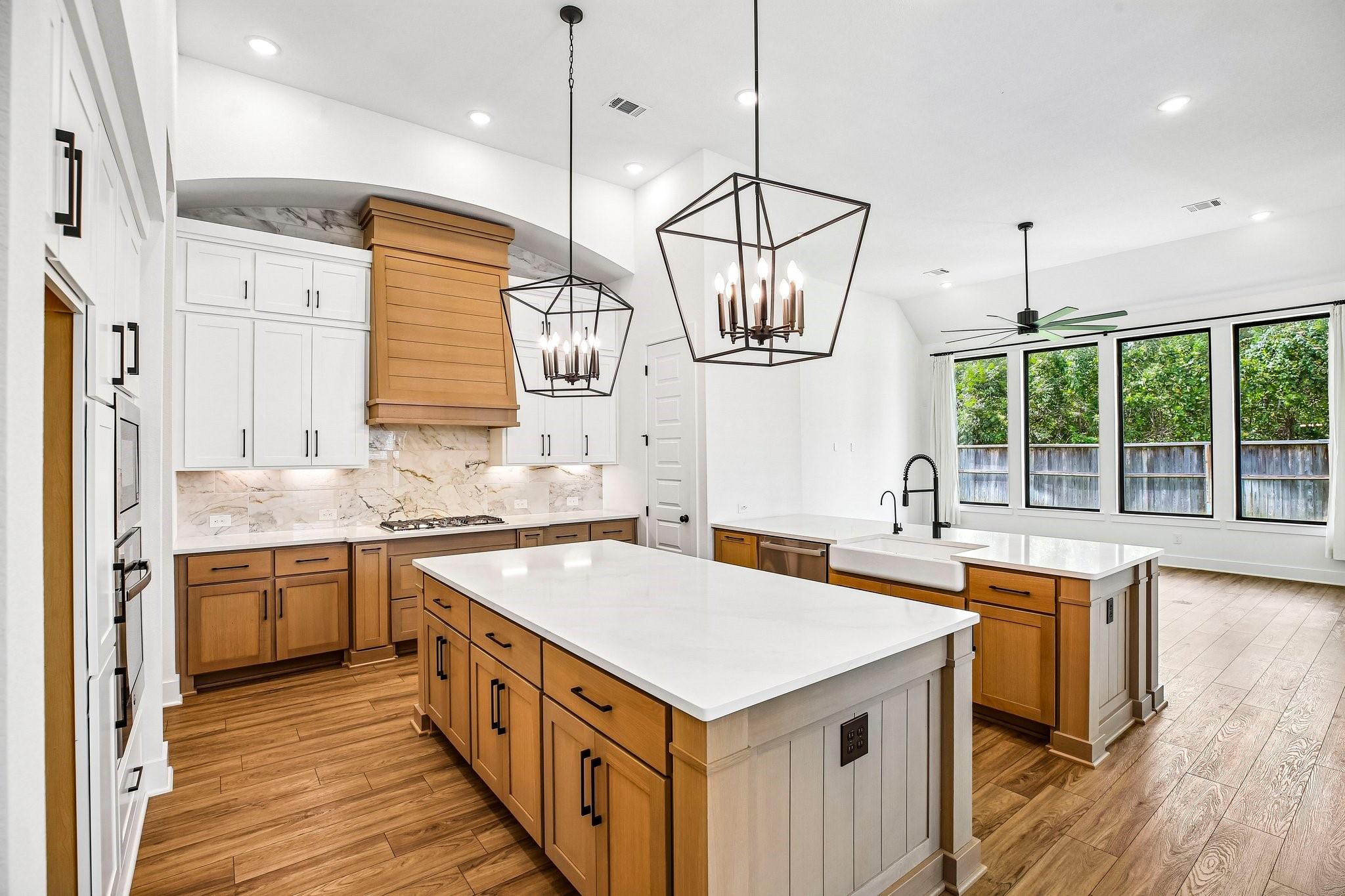 Spacious kitchen with two large islands, warm wood cabinetry, white counters, and wide windows overlooking trees.