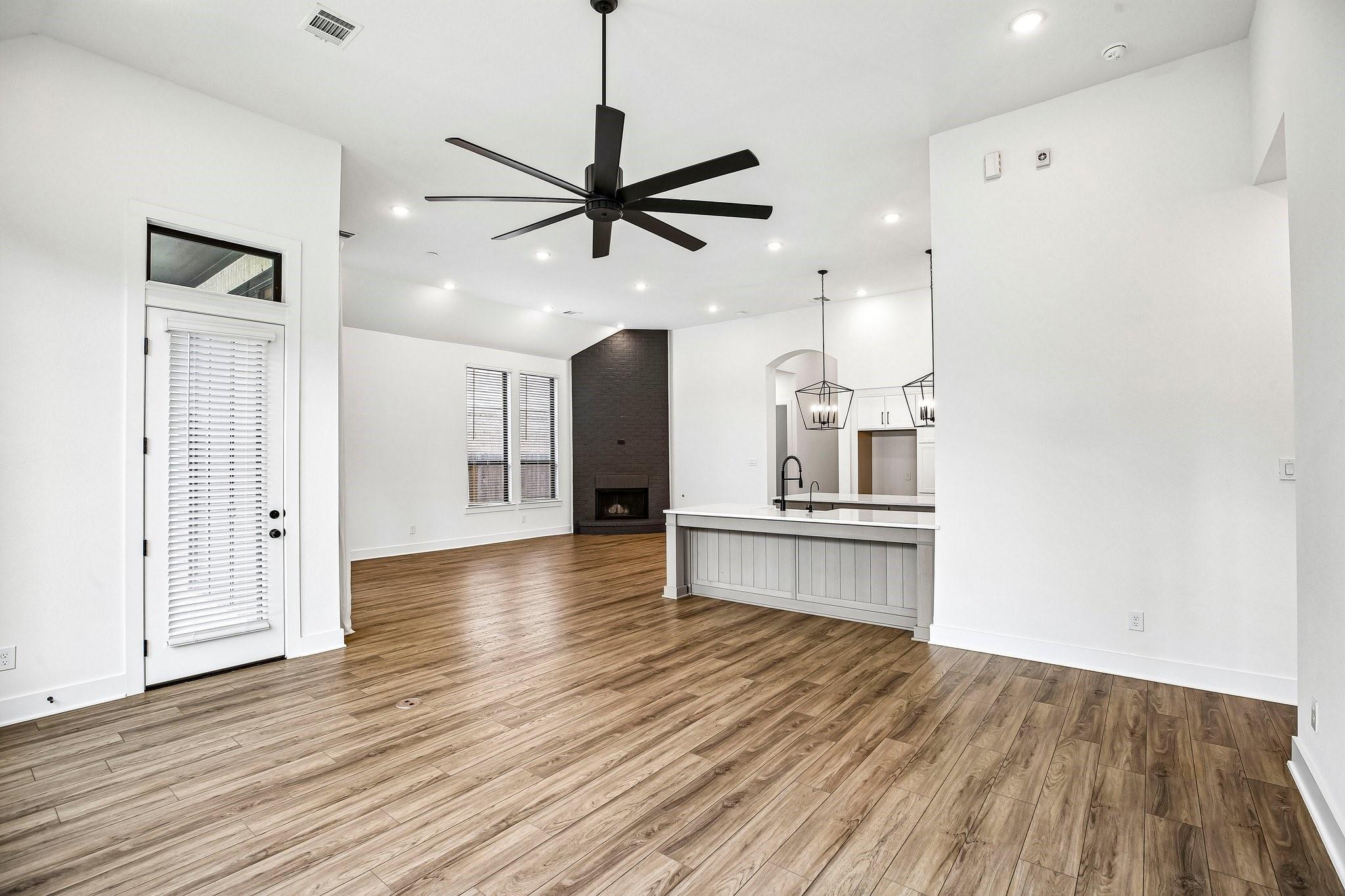 Vacant open-plan living room and kitchen with wood-look floors, a brick fireplace, and a large ceiling fan.