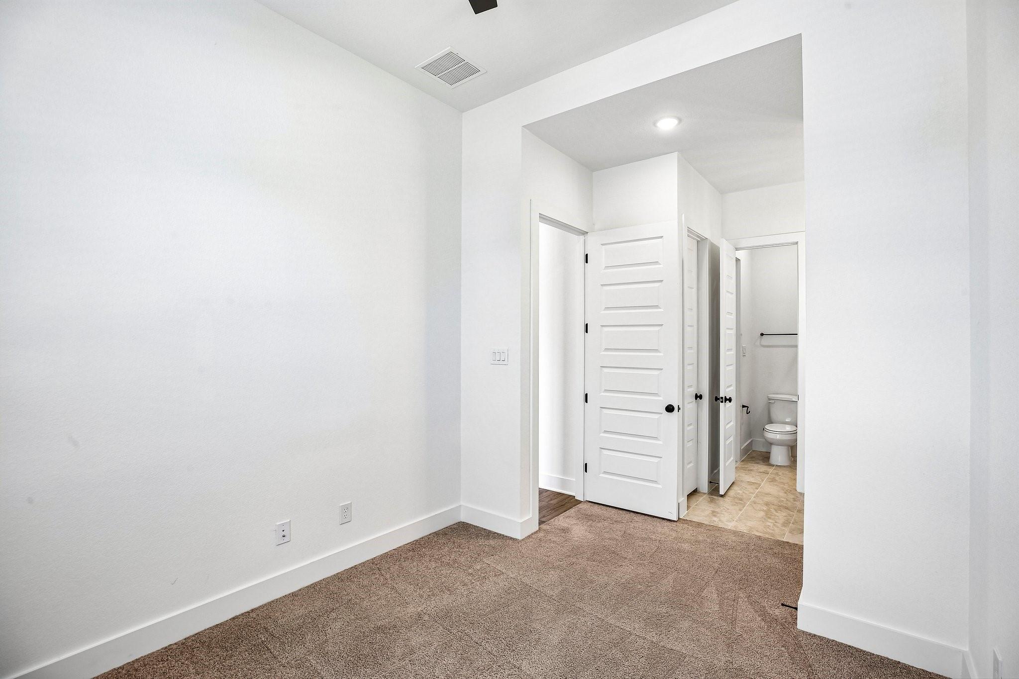 Empty carpeted bedroom with white paneled doors opening to a tiled bathroom.