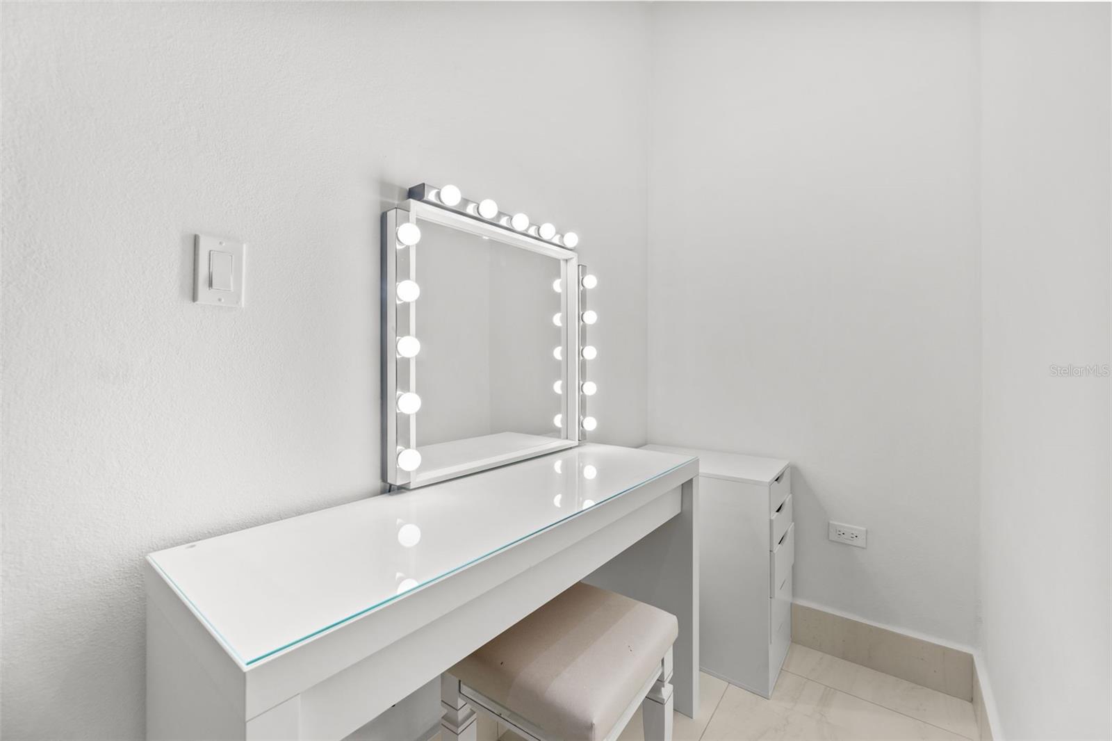 White dressing vanity with a padded stool and a mirror framed by bright bulbs.