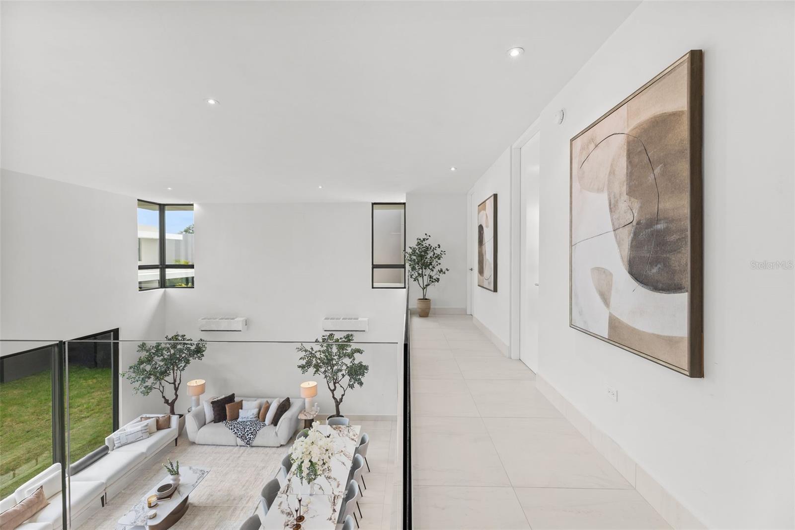 Upper-floor hallway overlooking a furnished living and dining area, with white tile floors and a glass railing.