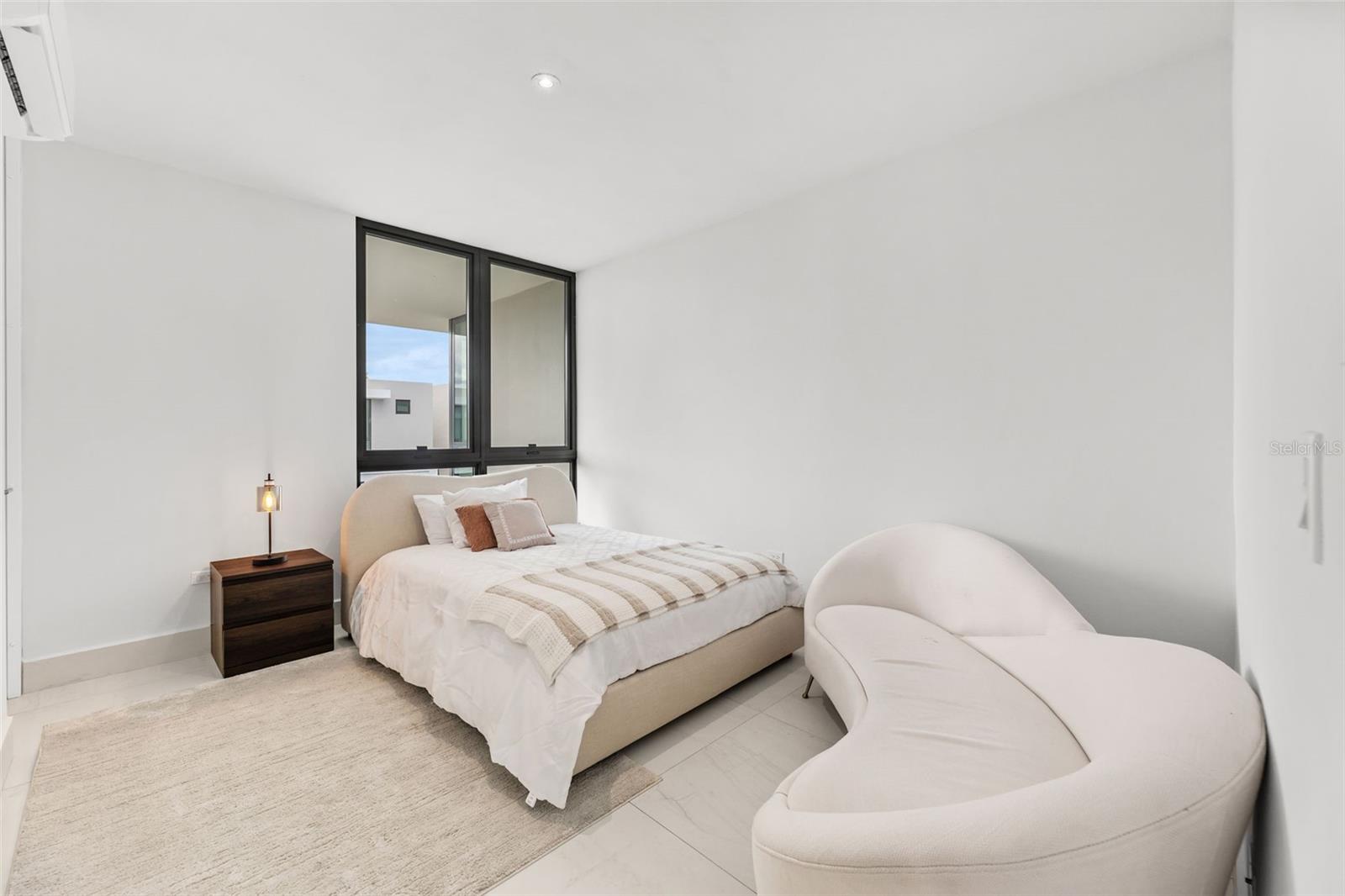 Furnished bedroom with a cream bed, curved lounge chair, and large black-framed window.