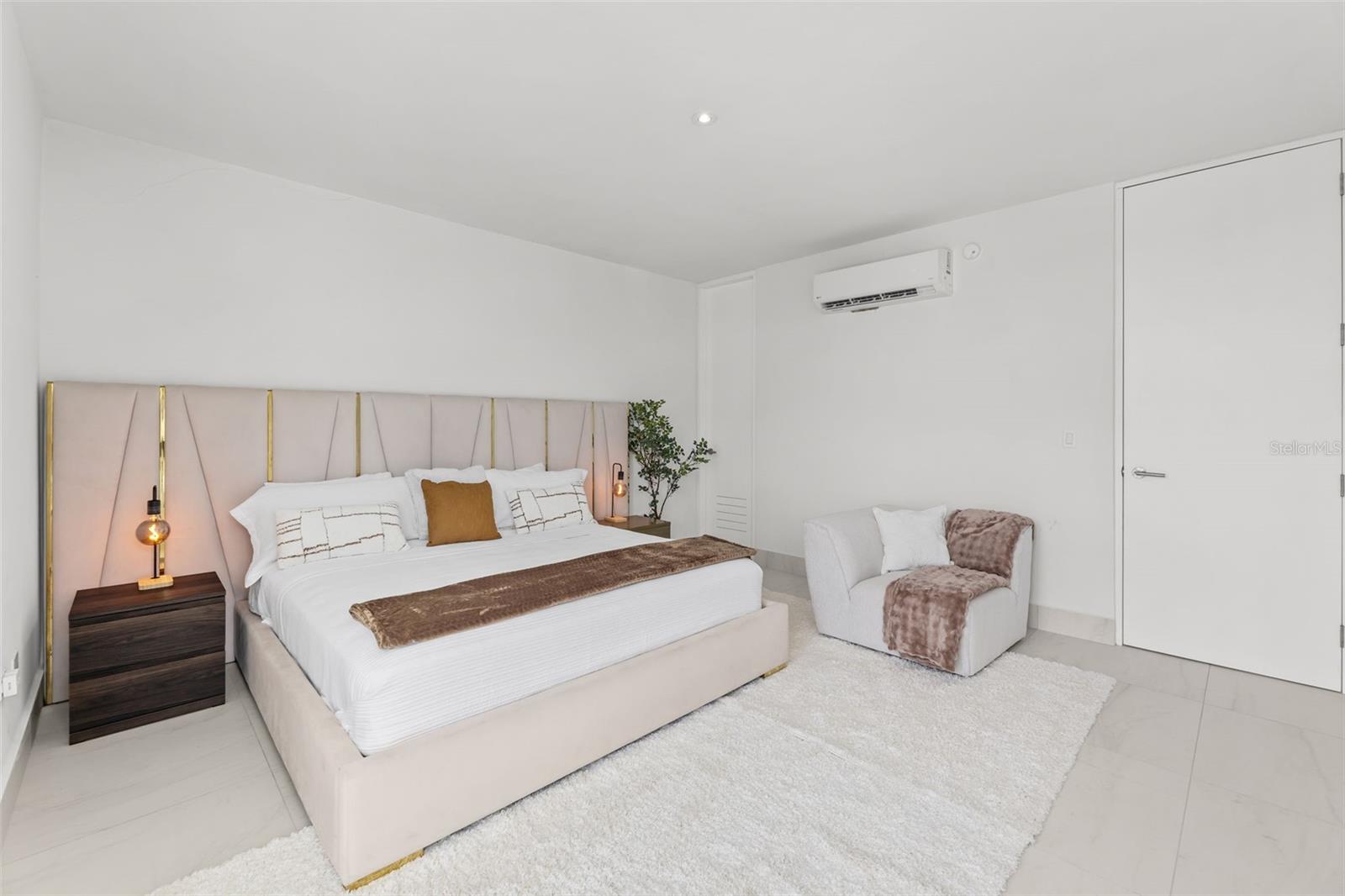 Staged bedroom with a blush upholstered bed, gold-accented headboard, and white armchair.