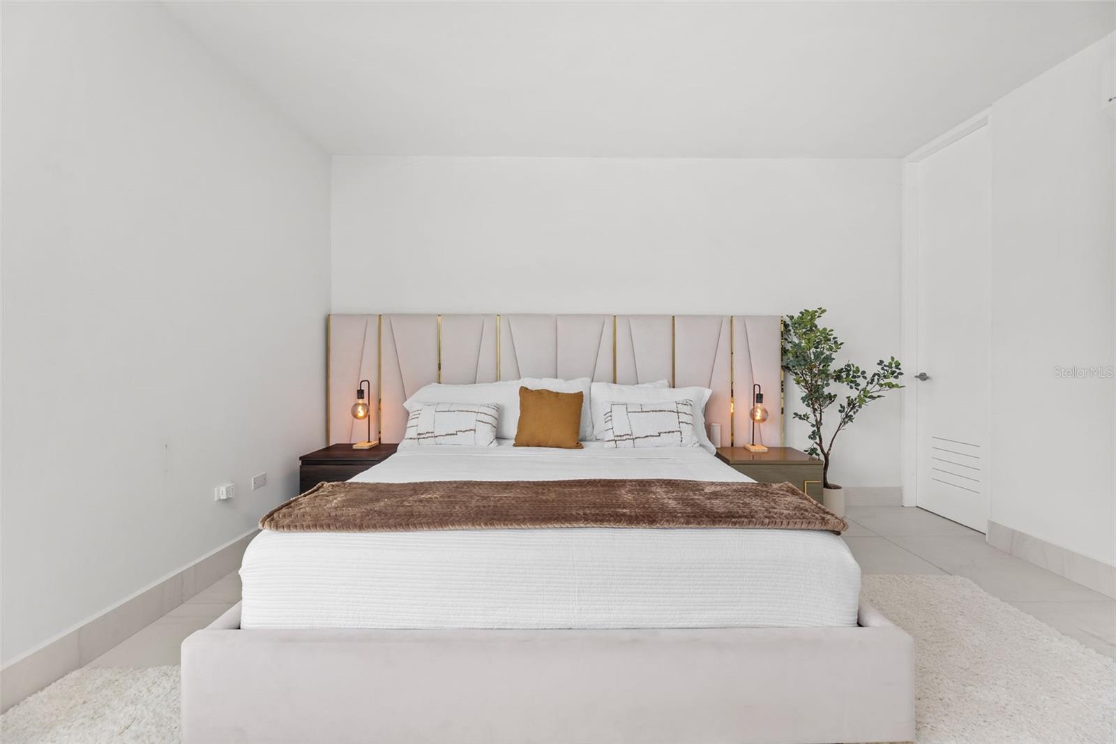 Staged bedroom with a large white bed, blush-and-gold headboard, bedside lamps, and a potted tree.