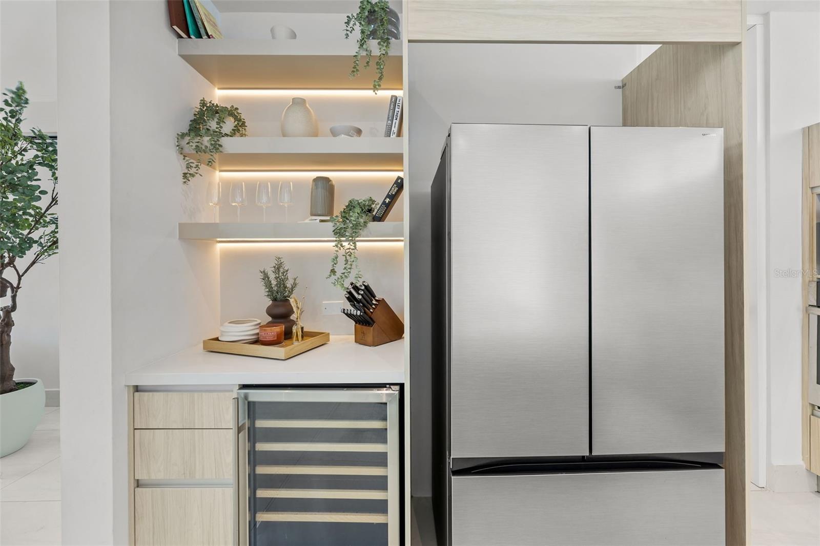 Modern kitchen with a stainless refrigerator, wine cooler, and illuminated display shelves.