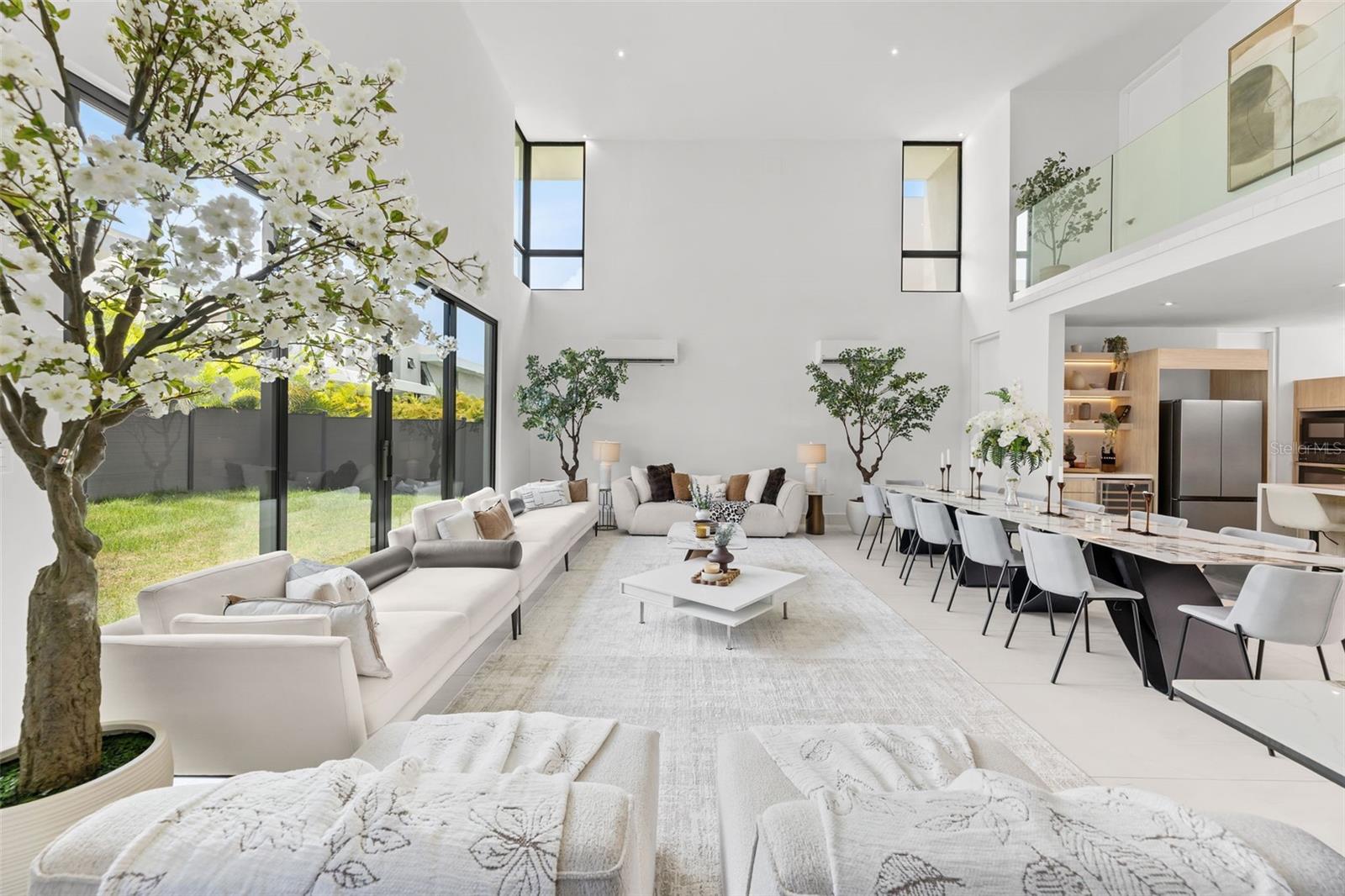Double-height living and dining room with white sofas, a long table, and flowering indoor trees.