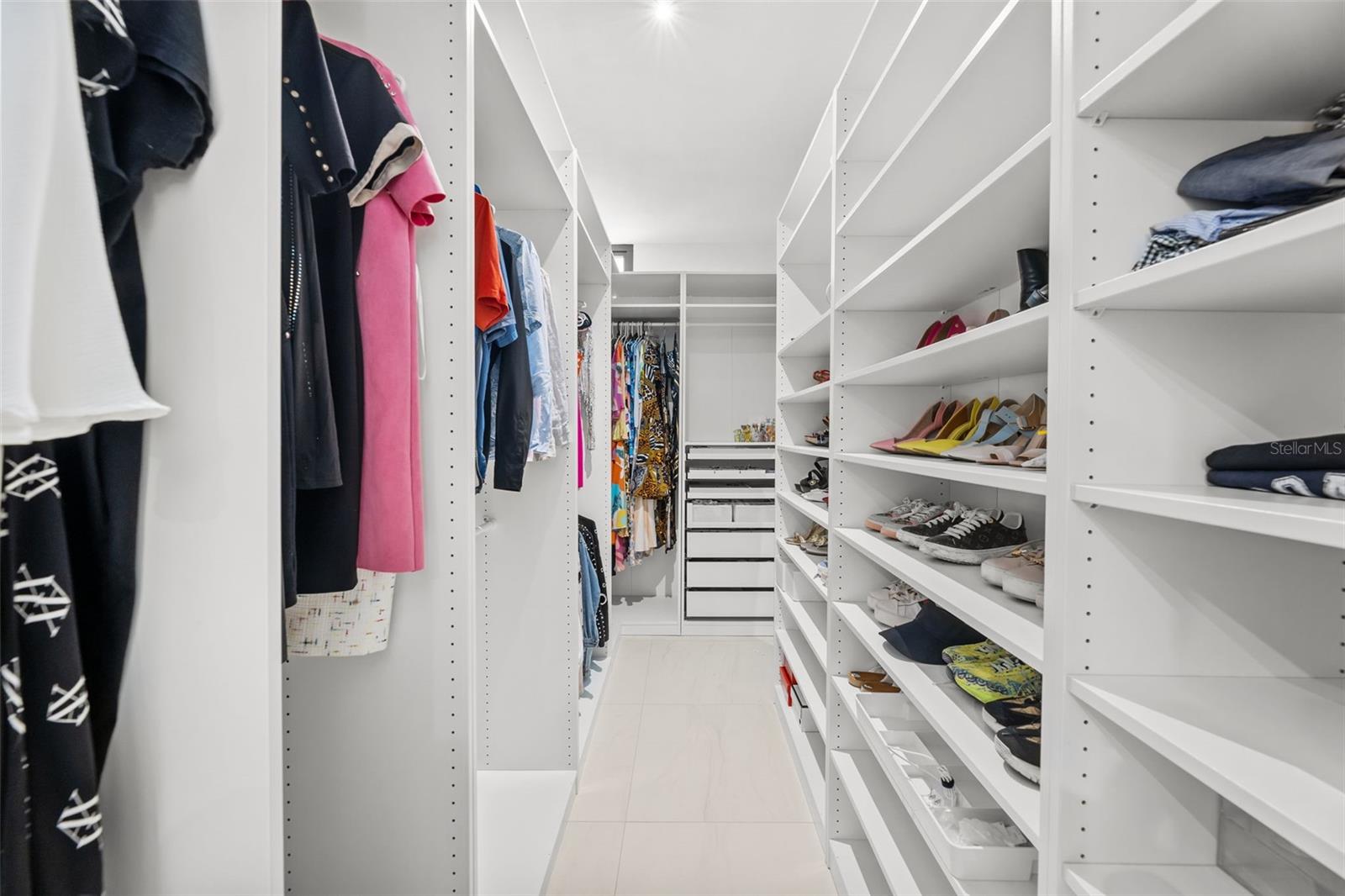 Walk-in closet with white built-in shelves, hanging clothes, and rows of shoes.