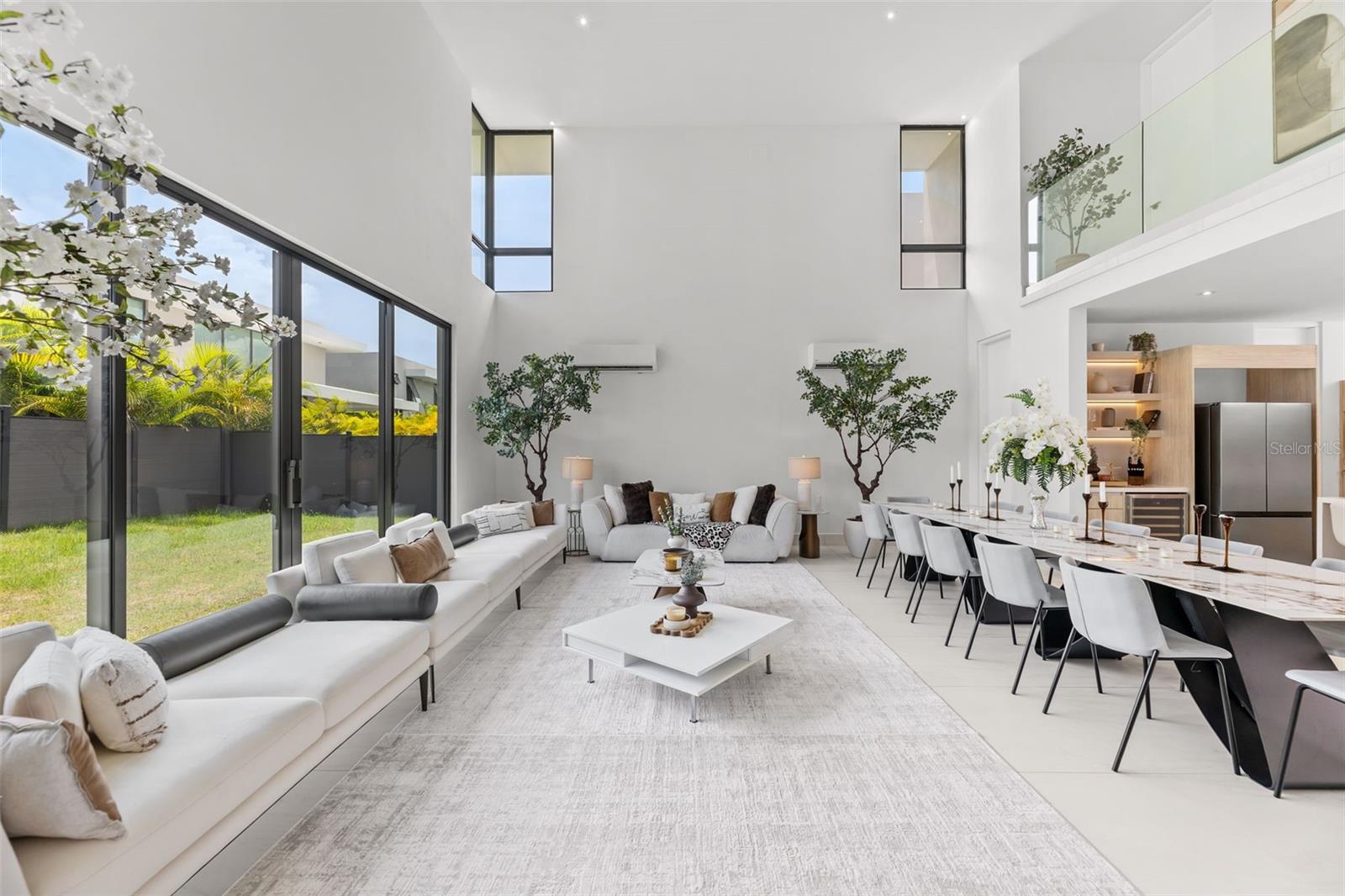 Double-height living room with white sofas, floor-to-ceiling windows, and a long dining table.