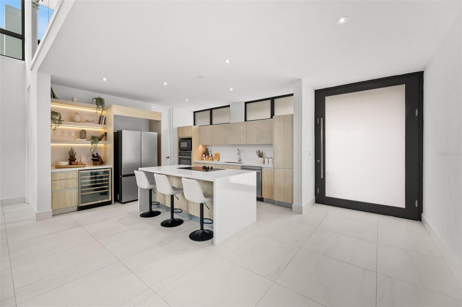 Modern kitchen with a large white island, light wood cabinets, stainless appliances, and illuminated display shelves.