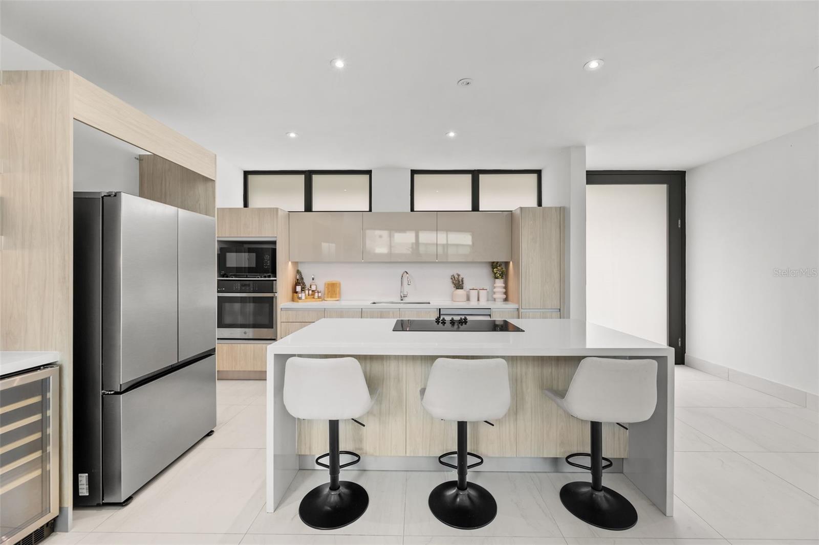 Modern kitchen with pale wood cabinets, a large island, three stools, and stainless-steel appliances.