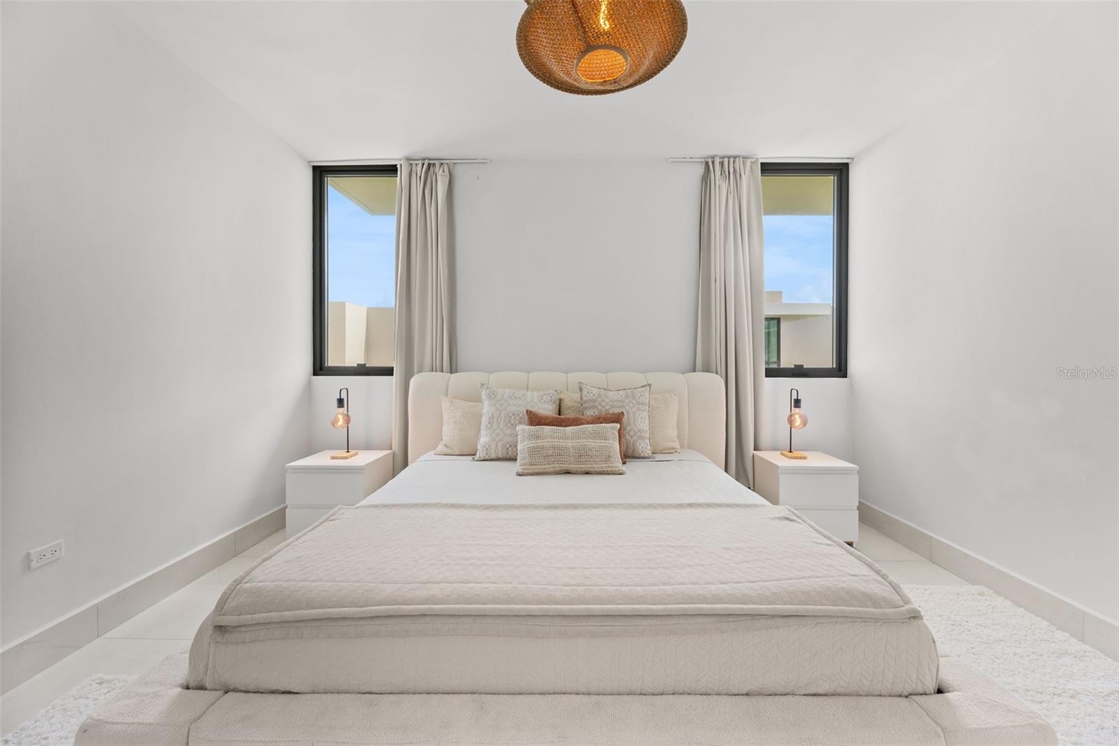 Staged bedroom with a cream bed, matching nightstands, and two curtained windows.