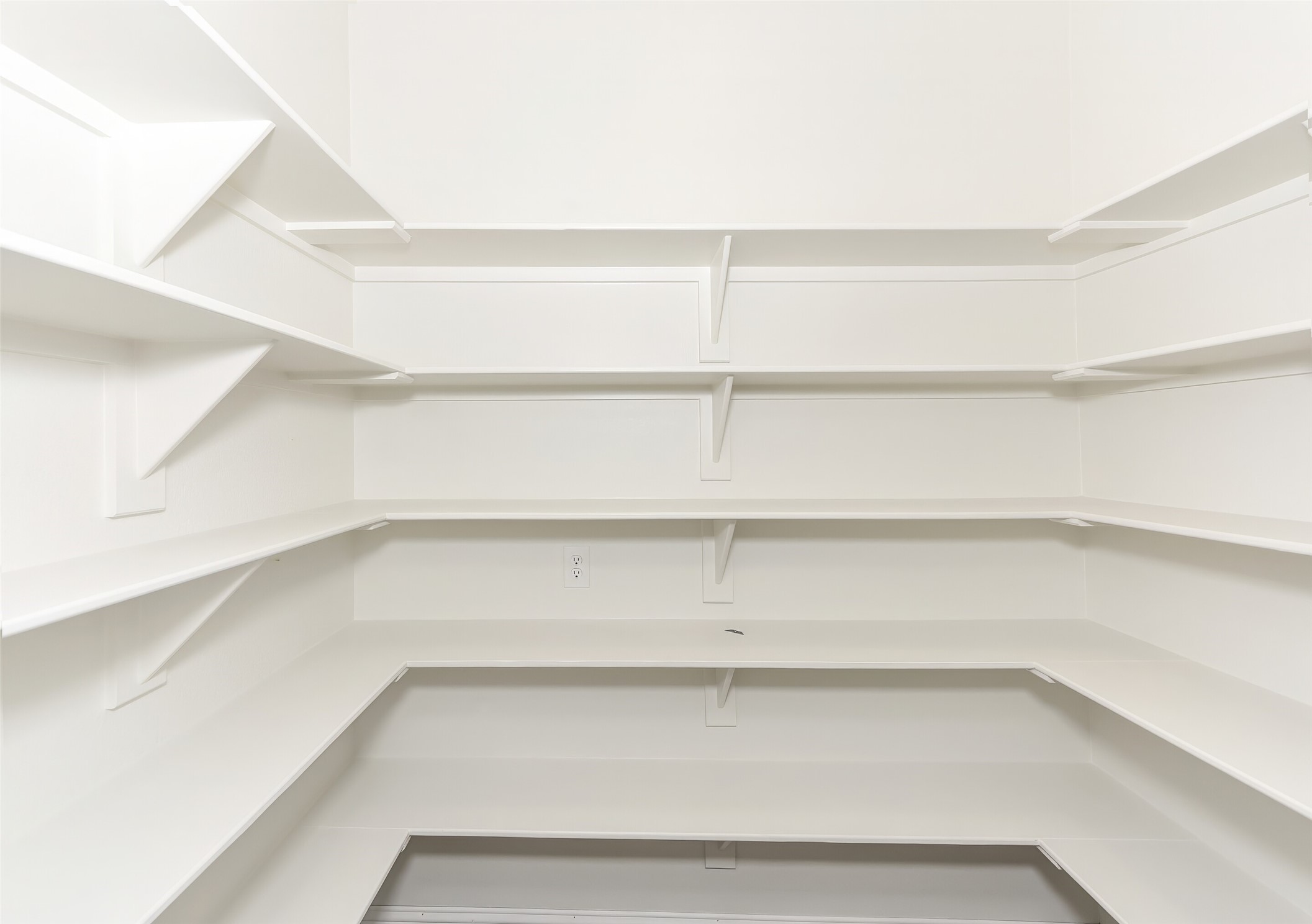 Empty walk-in pantry with white wraparound shelves and a wall outlet.