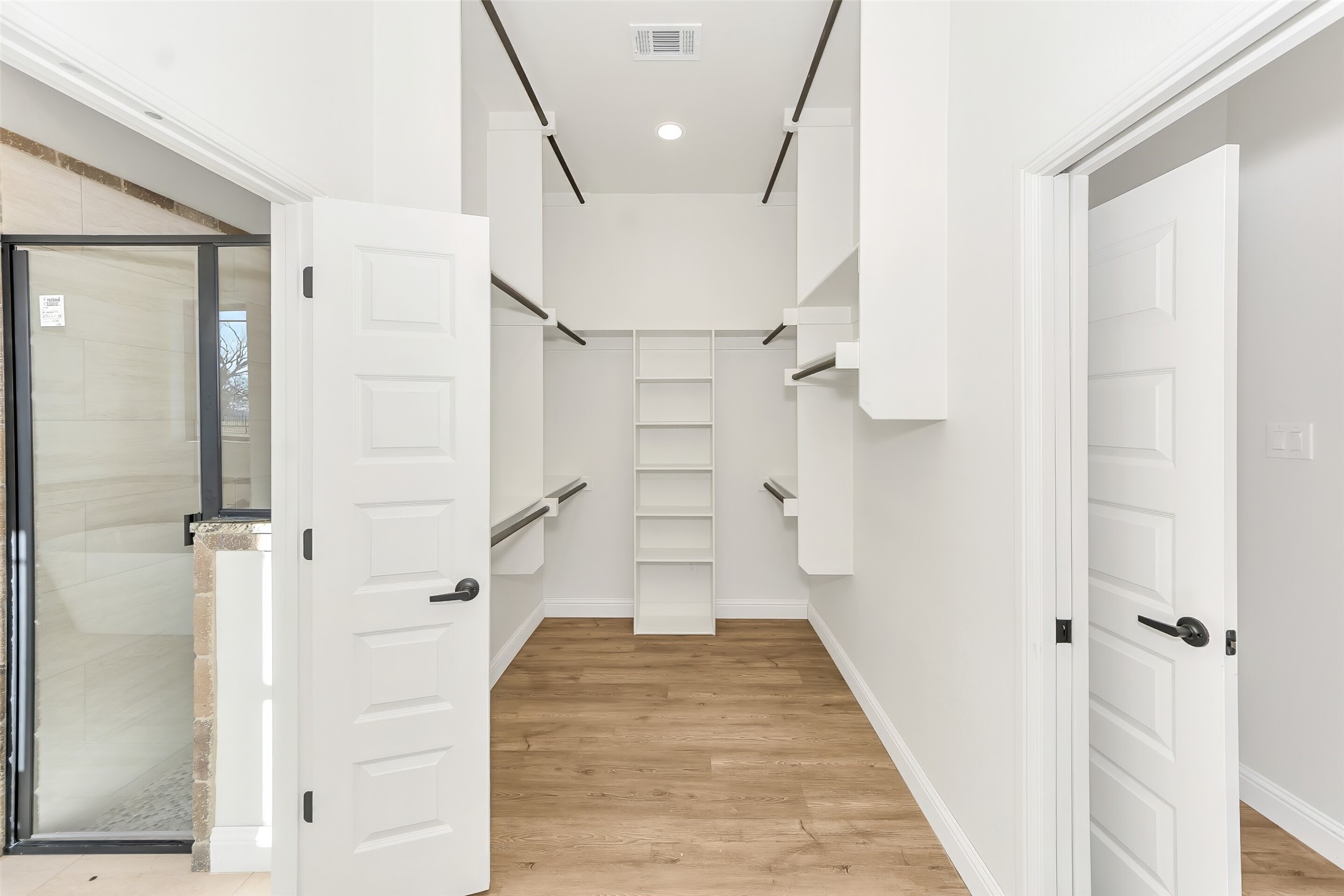 Walk-in closet with built-in shelving and wood flooring beside a glass-enclosed shower.