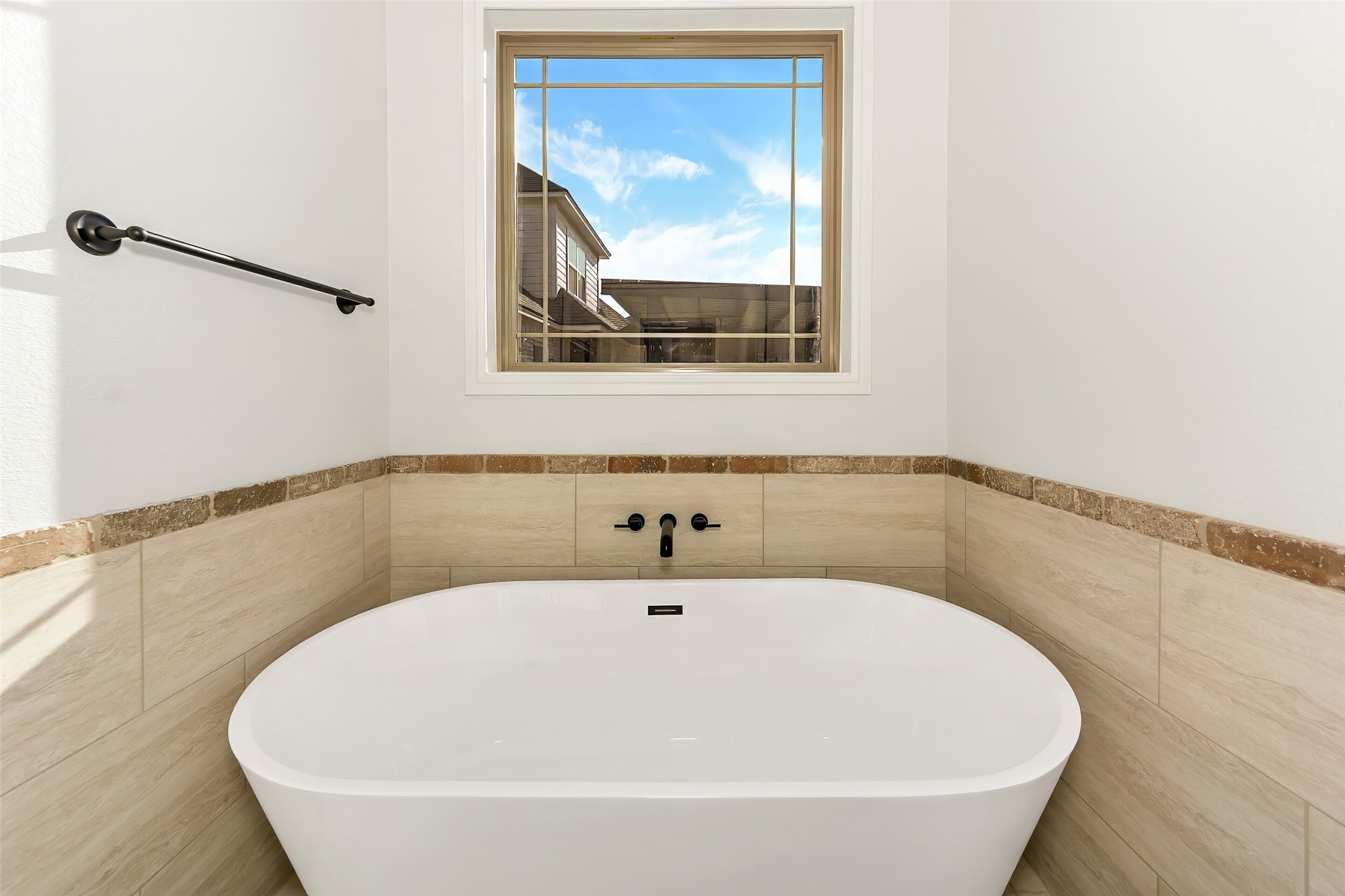 Freestanding bathtub beneath a large window, with beige tile and a wall-mounted grab bar.